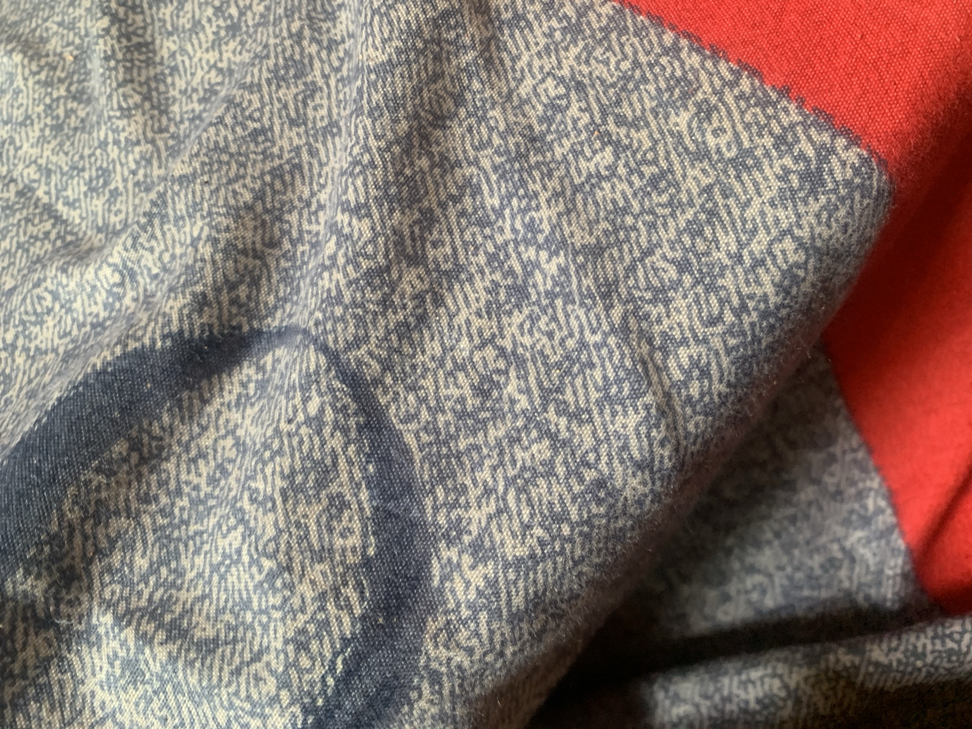 The image displays a close-up view of a textured fabric with a blue and white patterned print. The fabric is creased, suggesting it is folded or draped. A bright red fabric is visible along the bottom right edge of the image, appearing to be a contrasting trim or a separate piece of material. The lighting casts shadows that emphasize the texture and folds of the blue and white fabric. There are no discernible elements that place this image specifically in Jauro Yinu, Nigeria.
