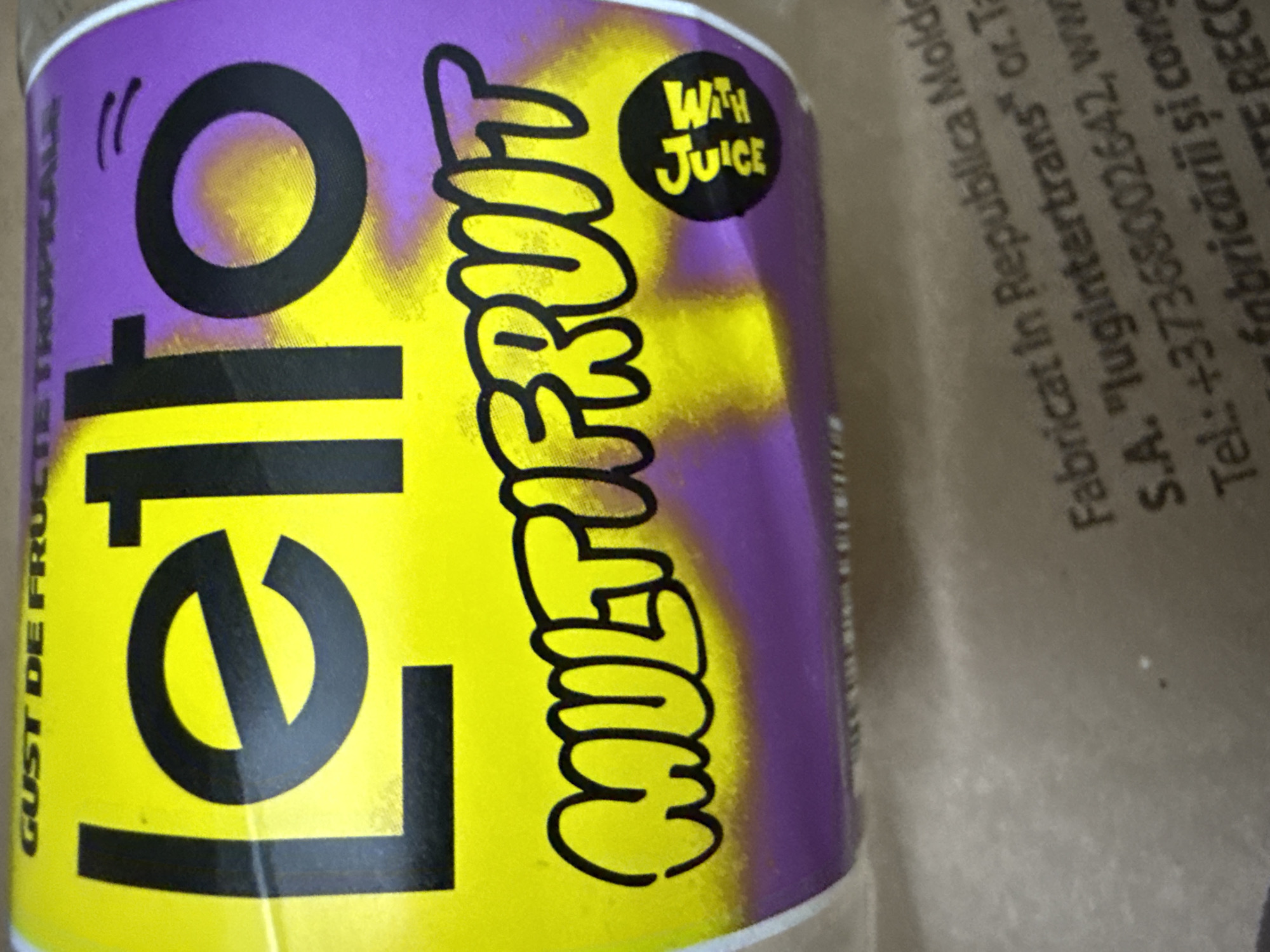 The image shows a close-up of a colorful beverage can, likely a juice or soda. The can features a vibrant design with a purple top fading into a bright yellow body. Prominently displayed on the can is the brand name "Lello" in large, bold black lettering. Below "Lello," the flavor "MULTIFRUIT" is written in a stylized, bubbly font. A small black circle with the text "WITH JUICE" is also visible. Vertical text on the left side of the can reads "GUST DE FRUC".
The right side of the image shows a portion of what appears to be a cardboard box or packaging, with text printed in black. This text is partially obscured and angled, but some phrases are legible, including "Fabricat în Republica Moldo" (Manufactured in the Republic of Moldova), "S.A. 'lugintertrans'", and a phone number "+37368002642". This suggests the beverage is produced in Moldova. The overall impression is of a commercial product, likely displayed for sale or as part of inventory. The lighting is somewhat dim, and the focus is on the can, with the background slightly blurred. There are no people or specific scenes depicted, only the product itself and its packaging information.