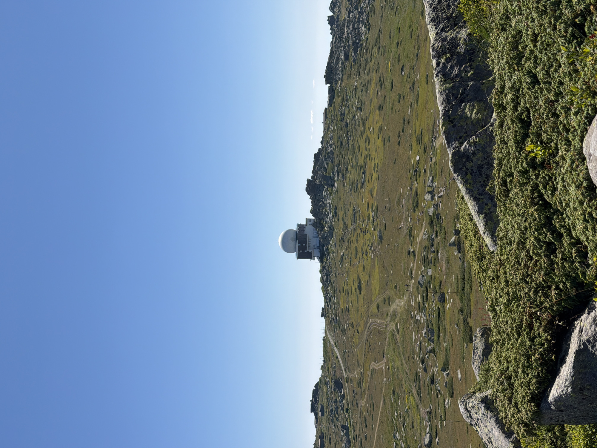 The image shows a clear blue sky over a rocky, mountainous terrain. In the middle ground, a white dome-shaped structure sits atop a building complex, likely a meteorological station or radar facility, located in the vicinity of Bistritsa, Bulgaria. The landscape is dotted with rocks of various sizes and patches of dry grass and low-lying green vegetation. Several dirt tracks wind through the terrain, suggesting pathways or roads. In the foreground, large rocks covered with green, low-growing plants are visible. The overall scene is dry and exposed, characteristic of a high-altitude environment.