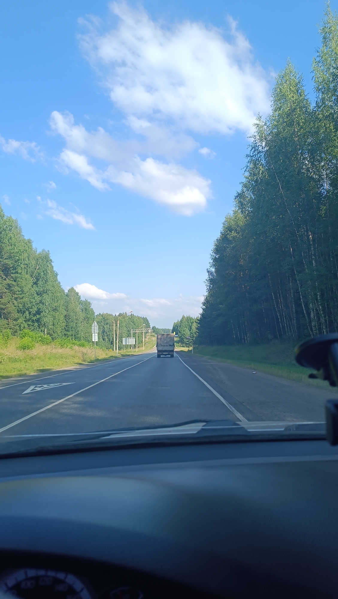 The image depicts a view from the interior of a vehicle driving on a two-lane road, likely in or near Garevaya, Russia, based on the context provided. The road is paved and lined with white markings. On the left side of the road, there is a grassy shoulder leading to a dense forest of green trees. On the right side of the road, a similar dense forest of trees, some appearing to be birch with white trunks, lines the road. The sky above is bright blue with scattered white clouds. In the distance, a dark-colored truck is traveling in the same direction as the camera vehicle. There are road signs visible on the left side of the road, including what appears to be a directional sign and a sign with a white arrow forming a triangle. Overhead structures, possibly for traffic signals or lighting, are also present in the distance. The dashboard of the vehicle is visible in the foreground, showing part of a speedometer. The overall scene suggests a drive through a rural, forested area on a sunny day.