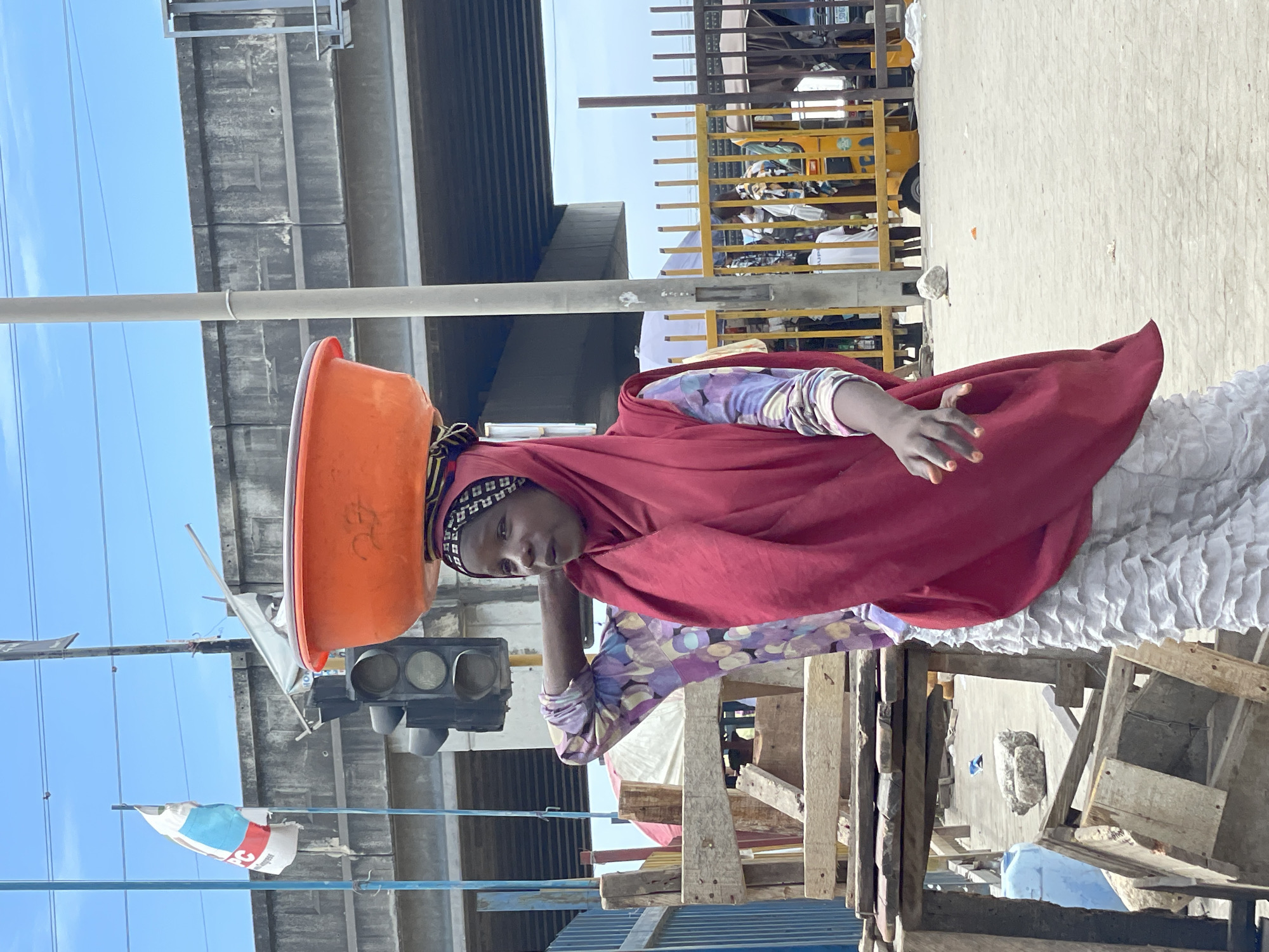 A woman with dark skin and a head covering is walking through a busy urban environment. She carries a large orange basin on her head. She is wearing a patterned long-sleeved shirt under a red shawl or scarf and appears to be in a crowded area with traffic and pedestrian activity.
The background includes a highway overpass, traffic lights, and a yellow vehicle with metal bars visible. There are flags on poles near the woman. The image composition suggests this is a public space, likely a street or market in Lekki, Nigeria. The presence of traffic infrastructure and what appears to be a public transportation vehicle indicates an active, populated setting.