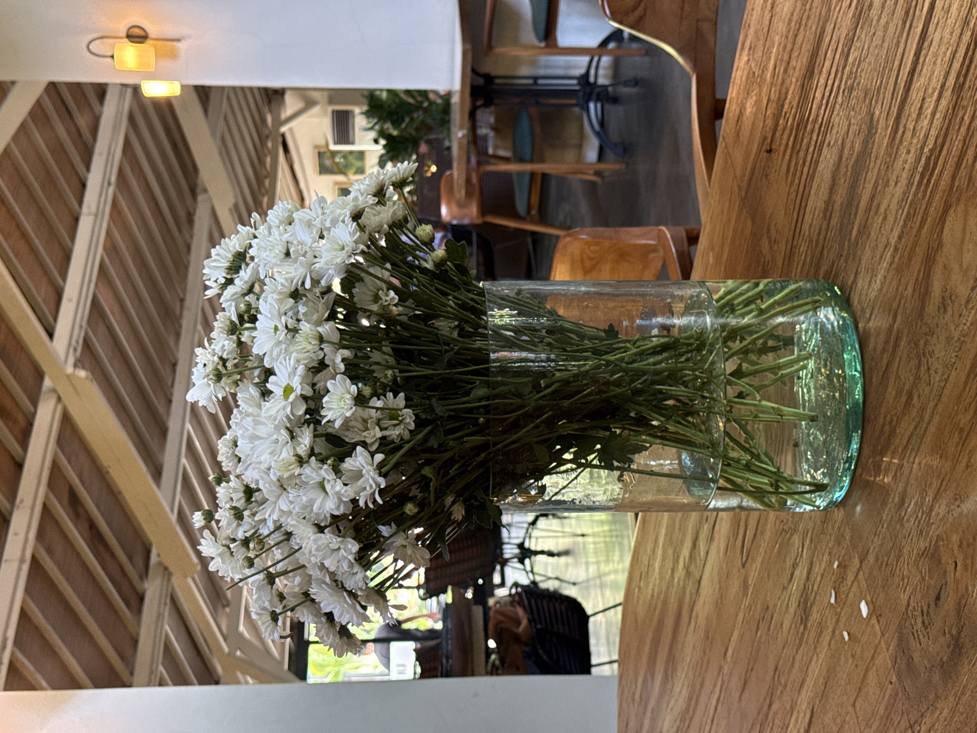 A static, eye-level indoor shot captures a vase of white flowers on a wooden table, likely in a cafe or restaurant in Ubud District, Indonesia. The image is oriented sideways, but described as if upright.
In the foreground, a clear glass vase, with a slight greenish tint at its base, holds a generous bouquet of small, white, daisy-like flowers, possibly chrysanthemums. Their green stems are visible through the water in the vase. The vase rests on a light brown, textured wooden table. To the right of the vase on the table, a few small, white, irregular-shaped specks are visible.
The background is softly blurred, focusing attention on the flowers. To the left, a white ceiling or wall panel features a warm yellow light fixture. Below this, a slatted wooden structure with diagonal beams forms part of the wall or ceiling. To the right, out-of-focus wooden chairs and tables are visible in a more dimly lit area, indicating other seating within the establishment. Some chairs have teal-colored upholstery. Reflections of the surroundings, including portions of the furniture and potentially an outdoor scene, are distortedly visible in the surface of the vase.
The overall lighting is bright and natural, suggesting it is daytime. The decor features natural wood tones, contributing to a warm and inviting atmosphere. There is no visible text.