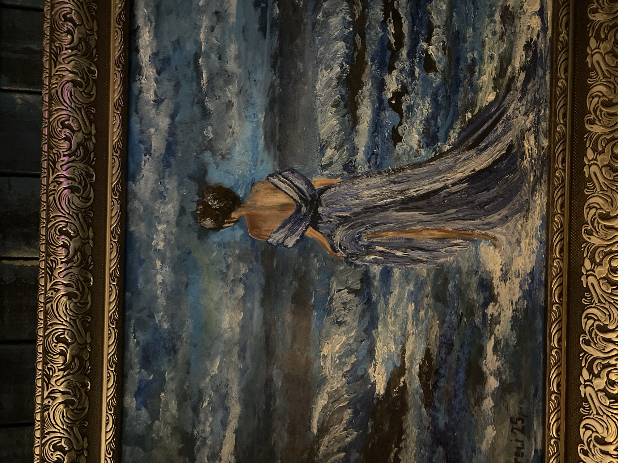 This is a framed oil painting depicting a woman in a blue dress standing at the edge of a turbulent sea. The woman is shown from the back, with her hair up, and her dress flowing. The sea is rendered with dramatic brushstrokes in shades of blue, white, and dark gray, suggesting rough waves and a cloudy sky. The painting is signed "orsci 25" in the lower left corner. The frame is ornate and golden. The image does not contain any information or visual cues that would allow for its location to be identified in the city of Poltava, Ukraine.
