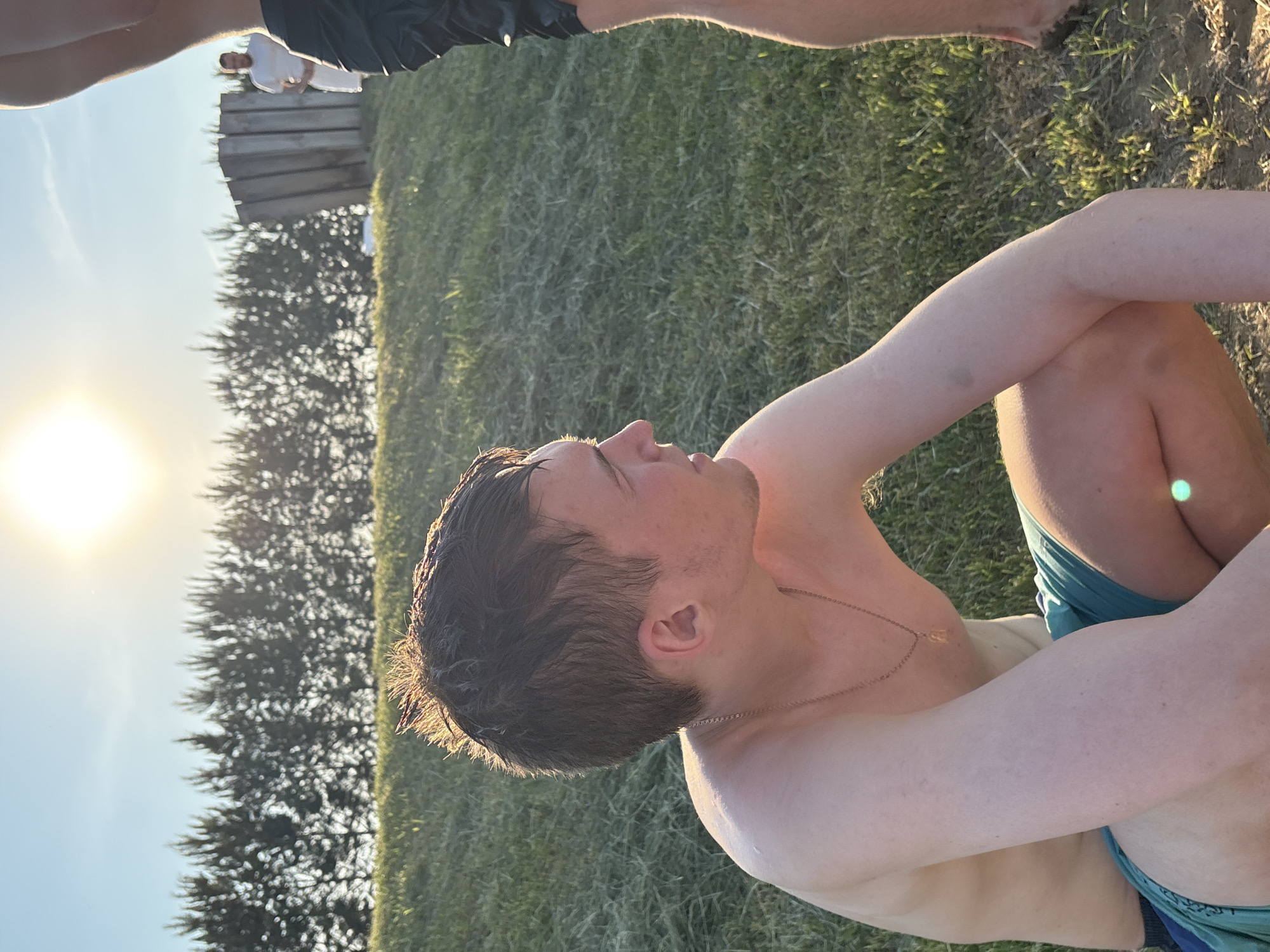 A forensic description of the image reveals an outdoor scene in Emel'yanovo, Russia, under bright, low-angle sunlight, indicative of either sunrise or sunset.
In the immediate foreground, a young Caucasian male, shirtless and wearing blue/green swim trunks, is captured in a crouched or seated position. His hair is dark brown, wet, and spiky, with individual strands reflecting the warm sunlight. He wears a thin, gold-colored chain with a small pendant around his neck. His skin exhibits a fair complexion with some reddish tones from sun exposure. His gaze is directed towards the right side of the frame, and his expression is neutral. His body, particularly his upper back and arms, shows droplets of water.
To the right, partially cropped, are the lower torso and legs of another individual, wearing dark, wet shorts. Only one leg is clearly visible, positioned towards the bottom right edge of the frame.
In the mid-ground, on a slight elevation covered with dry grass, stands a line of evenly spaced coniferous trees, somewhat out of focus. To the right of these trees, a rustic wooden structure, constructed from vertical planks, is visible. Adjacent to this structure, a third male figure stands, wearing a white t-shirt and white shorts or a white towel wrapped around his waist. This individual has facial hair.
The ground consists of dry grass and patches of bare earth. The sky is clear and light blue, with a prominent, bright sun disc located in the upper center-right, serving as the primary light source. No direct interactions between the individuals are discernible.