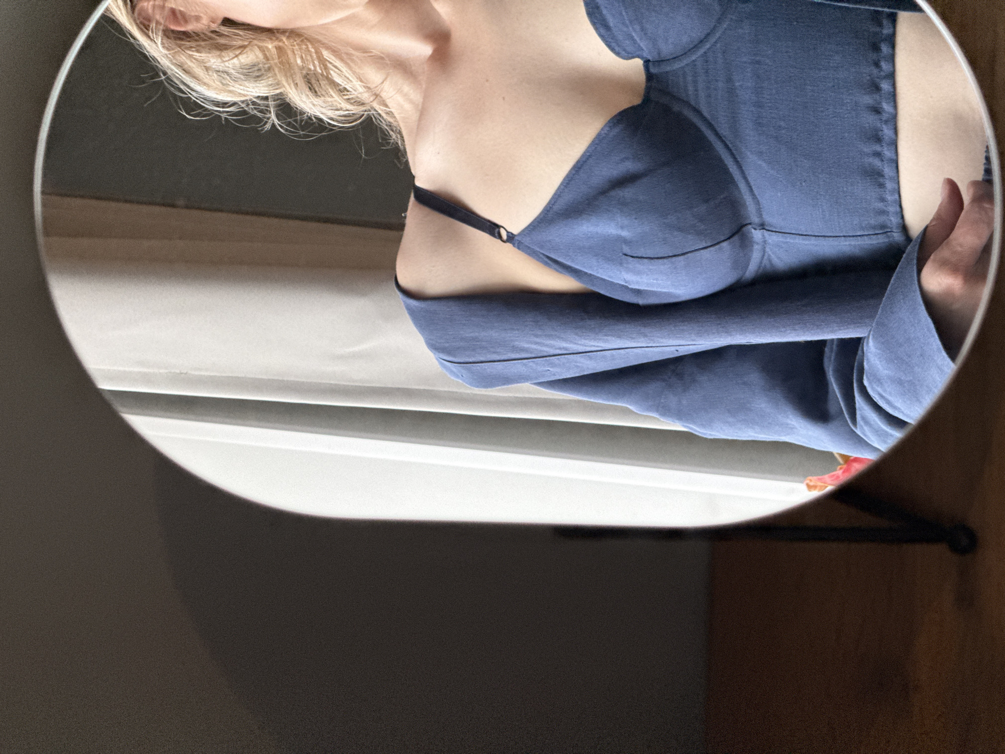 A woman with blonde hair is reflected in a round mirror. She is wearing a blue top, and her hand is visible on the right side of the mirror. The background includes a window with slats and a dark gray wall. The mirror hangs on a dark colored hanger that is attached to a wooden piece. There is a pink flower that is visible near the right side of the mirror. There is no evidence to indicate that the photograph was taken in Chaikovsky, Russia.