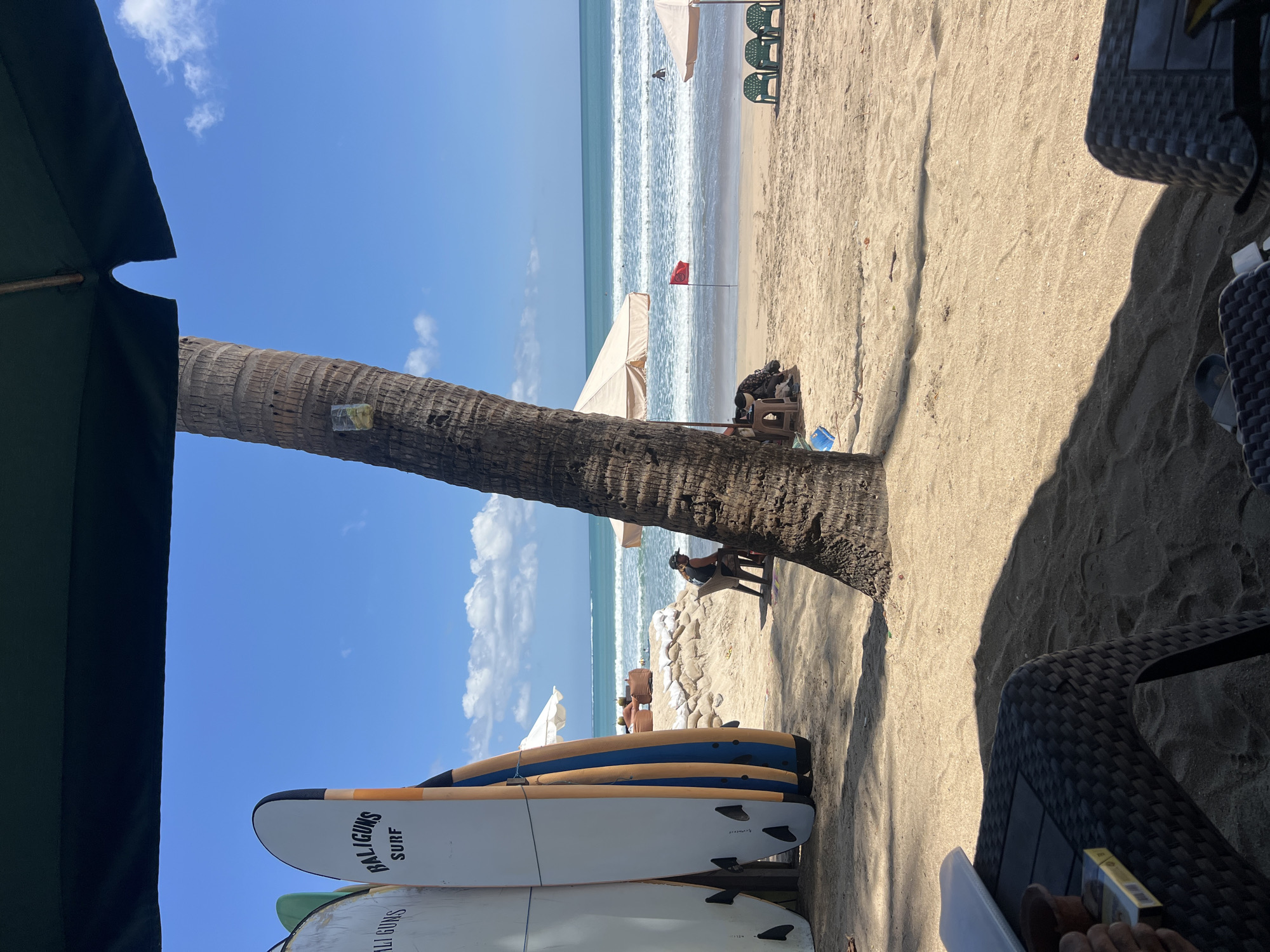 A beach scene in Kuta, Indonesia, features a foreground with a black woven table and chairs casting shadows. A pack of cigarettes sits on the table. Behind this, a thick palm tree trunk dominates the mid-ground, with a clear plastic cup attached to it. Leaning against a support structure are several surfboards, two of which are white with orange and blue accents and "BALIGUNS SURF" branding. Further back, a sandy beach stretches to the ocean, where waves are breaking. A red flag is planted in the sand near the water's edge. Two green beach chairs are visible on the sand. People are sparsely present, with one person sitting on a chair near the palm tree and others visible in the water or distant seating areas. The sky is bright blue with scattered white clouds. Beach umbrellas are present, one a neutral beige shade, and the other a darker green canopy overhead.