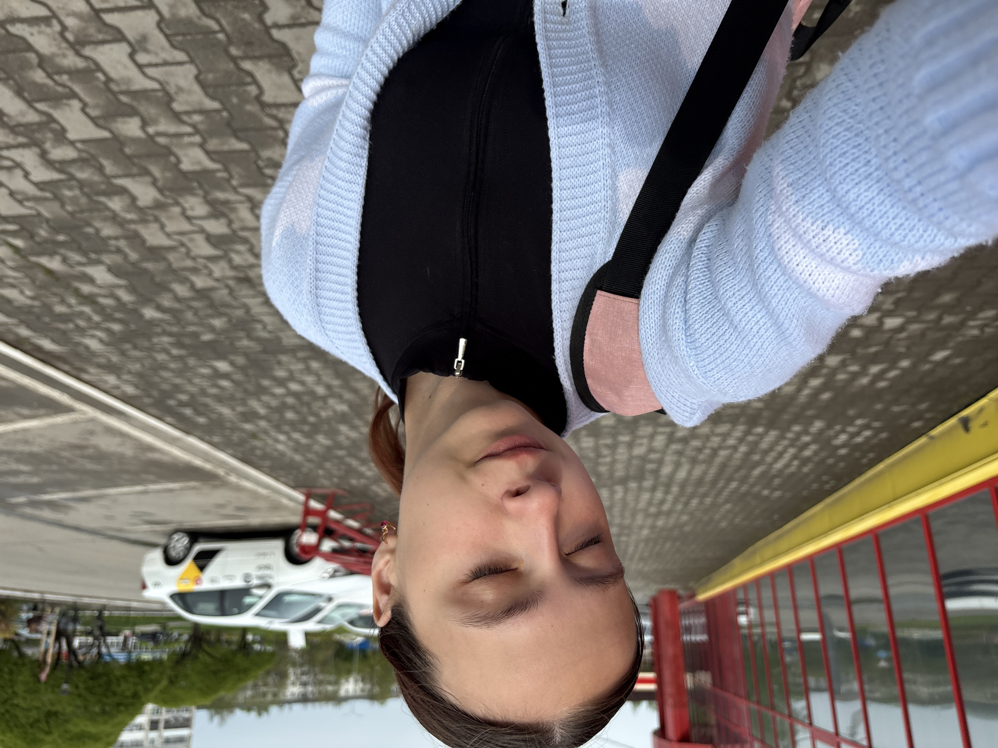 The image is a low-angle selfie of a young woman outdoors. She has fair skin, dark hair pulled back, and is wearing a black zippered top under a light blue cardigan. A black strap with a pink accent is visible across her body, likely from a bag.
The background shows a paved area made of interlocking blocks, leading to a red and yellow railing. Beyond the railing, a body of water reflects the sky and some greenery.
A white car is visible in the background, with a red metal frame and an identifiable yellow triangle with black markings. This car might be part of a driving school or training area. The presence of other distant parked cars, buildings, and trees indicates an urban or suburban setting.
Given the prompt mentioning the city of Chaikovsky, Russia, it's plausible the location is a riverside area or training center within that city.