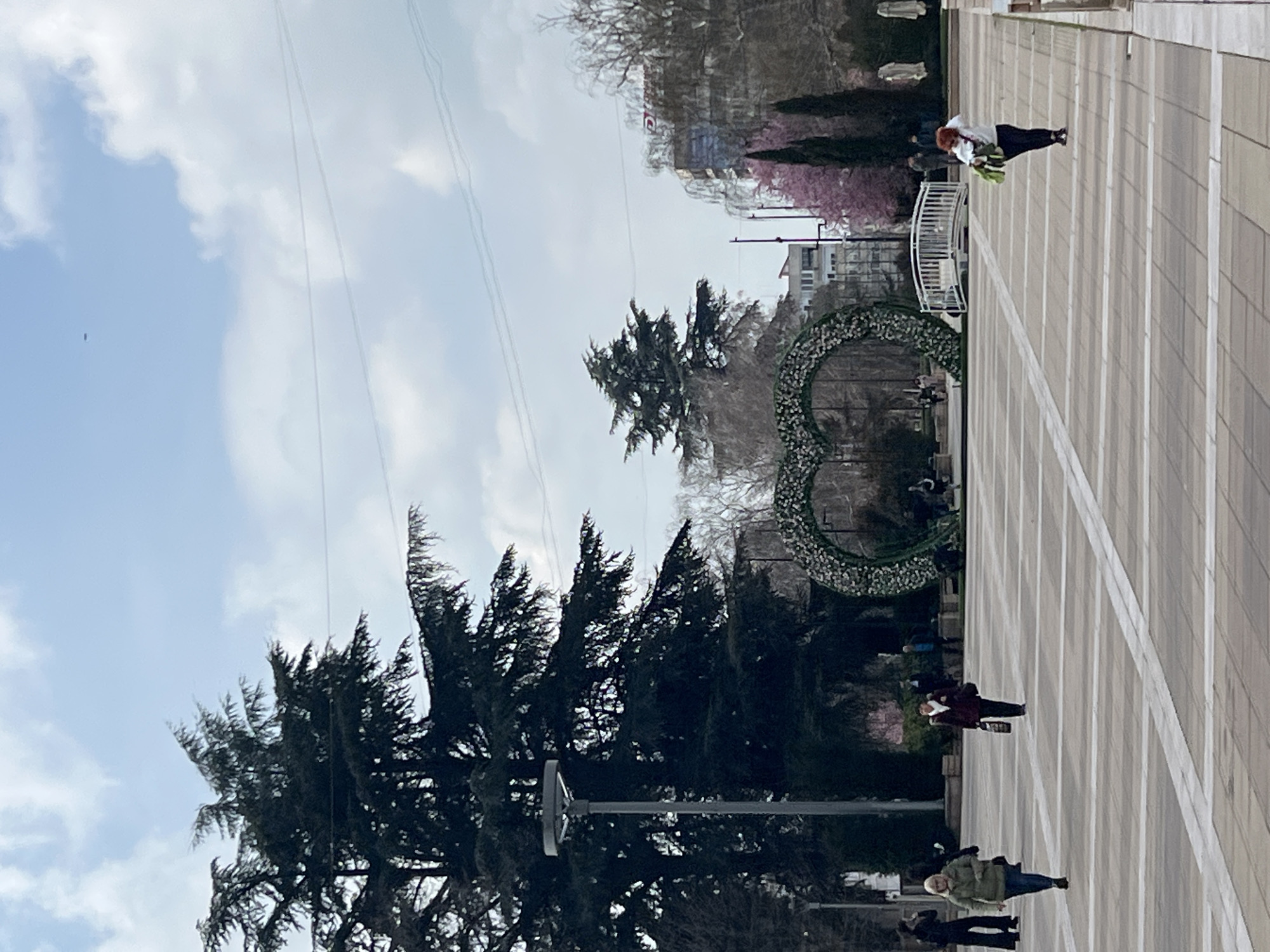 The image shows a public square in Yambol, Bulgaria. Several people are visible walking across the paved area. A large, heart-shaped floral arrangement stands near a white metal fence and flowering trees. Tall evergreen trees line part of the square. Overhead power lines are visible against a partly cloudy sky. Buildings can be seen in the background.