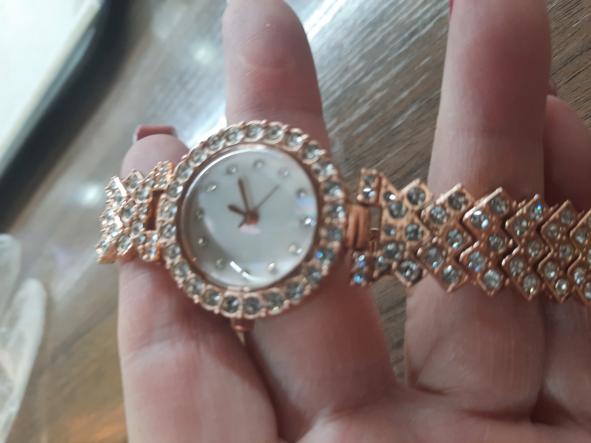 A close-up image shows a person's hand holding a highly decorative wristwatch against a blurred light wooden background. The watch features a round face with a rose gold-toned bezel, both heavily encrusted with numerous small, clear, sparkling crystals. The watch face is white or light silver with subtle crystal markers for hours and rose gold-toned hour and minute hands. The watch band is also rose gold-toned and designed with a geometric pattern, each segment of which is filled with multiple clear crystals, creating a continuous sparkling effect. The hand holding the watch has light-toned skin, and the fingernail of the index finger has light pink polish. The watch is the primary subject in sharp focus, highlighting its intricate, crystal-studded design. No specific activity is depicted other than holding the watch, possibly for display. The time of day and exact location are unknown. There is no visible text in the image.