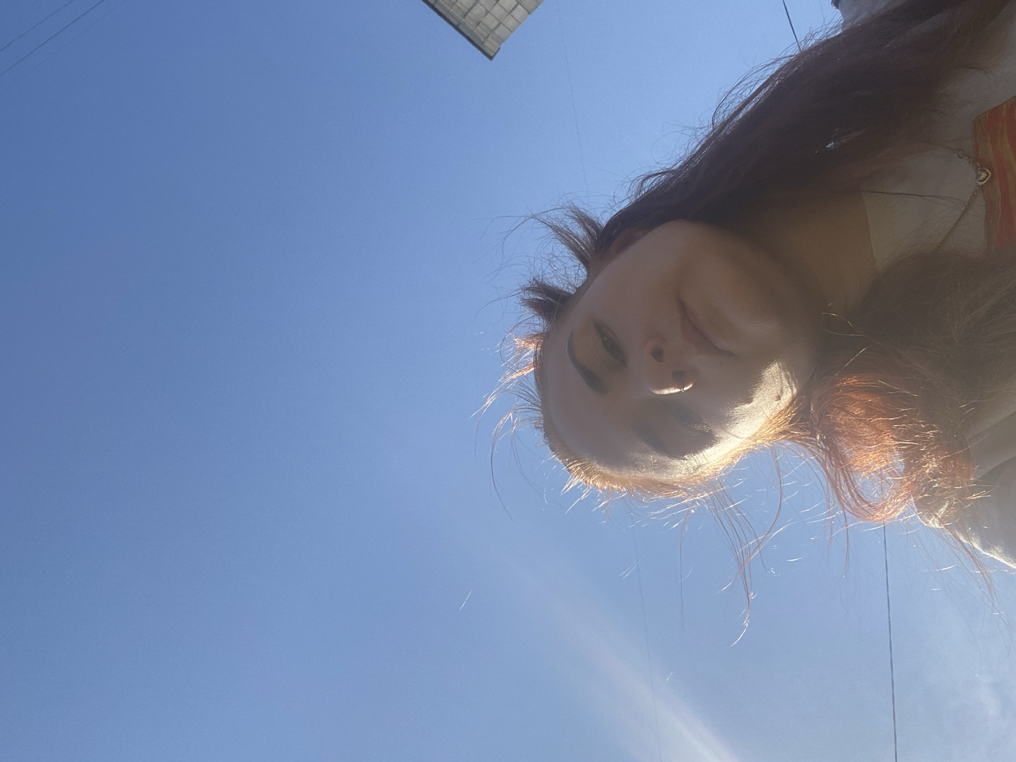 A low-angle shot shows a person's face against a bright blue sky. The person's head is tilted, with the nose pointing upwards. The person has dark hair with some reddish highlights. The person appears to have a piercing in their nose. A building with a grid-like structure is visible in the top center of the frame. Powerlines are visible. Given the prompt, it's reasonable to assume this image was taken in Chaikovsky, Russia.