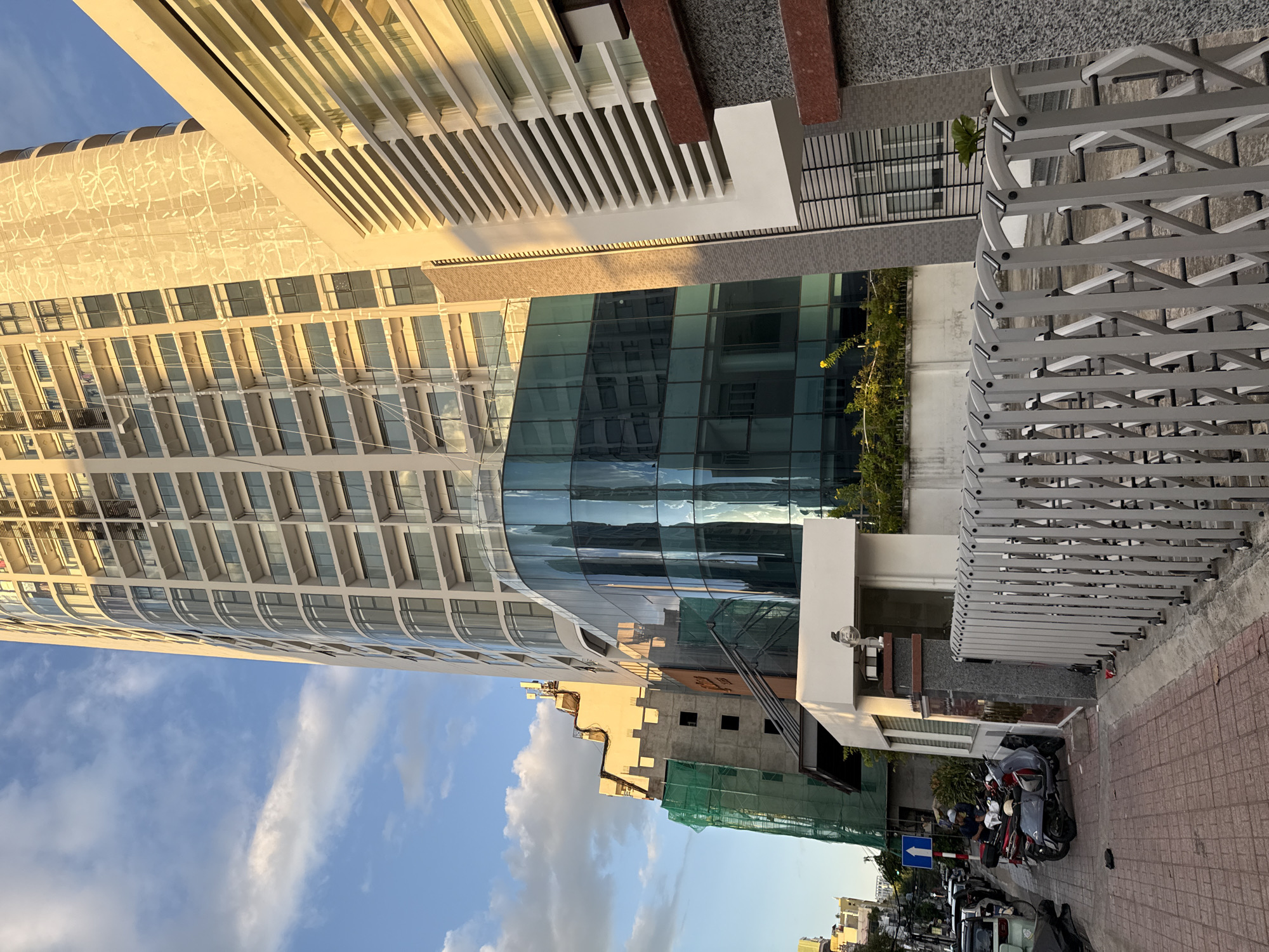 The image shows a street scene in Nha Trang, Vietnam, dominated by a tall, modern skyscraper under construction. The skyscraper has a curved facade made of glass and numerous balconies. To the left of the skyscraper, another building is also under construction, covered in green netting. In the foreground, a paved sidewalk runs alongside the street. Several parked motorcycles are visible on the sidewalk. A silver retractable gate is prominent in the lower right portion of the image. The sky is partly cloudy with a clear blue sky visible. The lighting suggests it is either early morning or late afternoon.