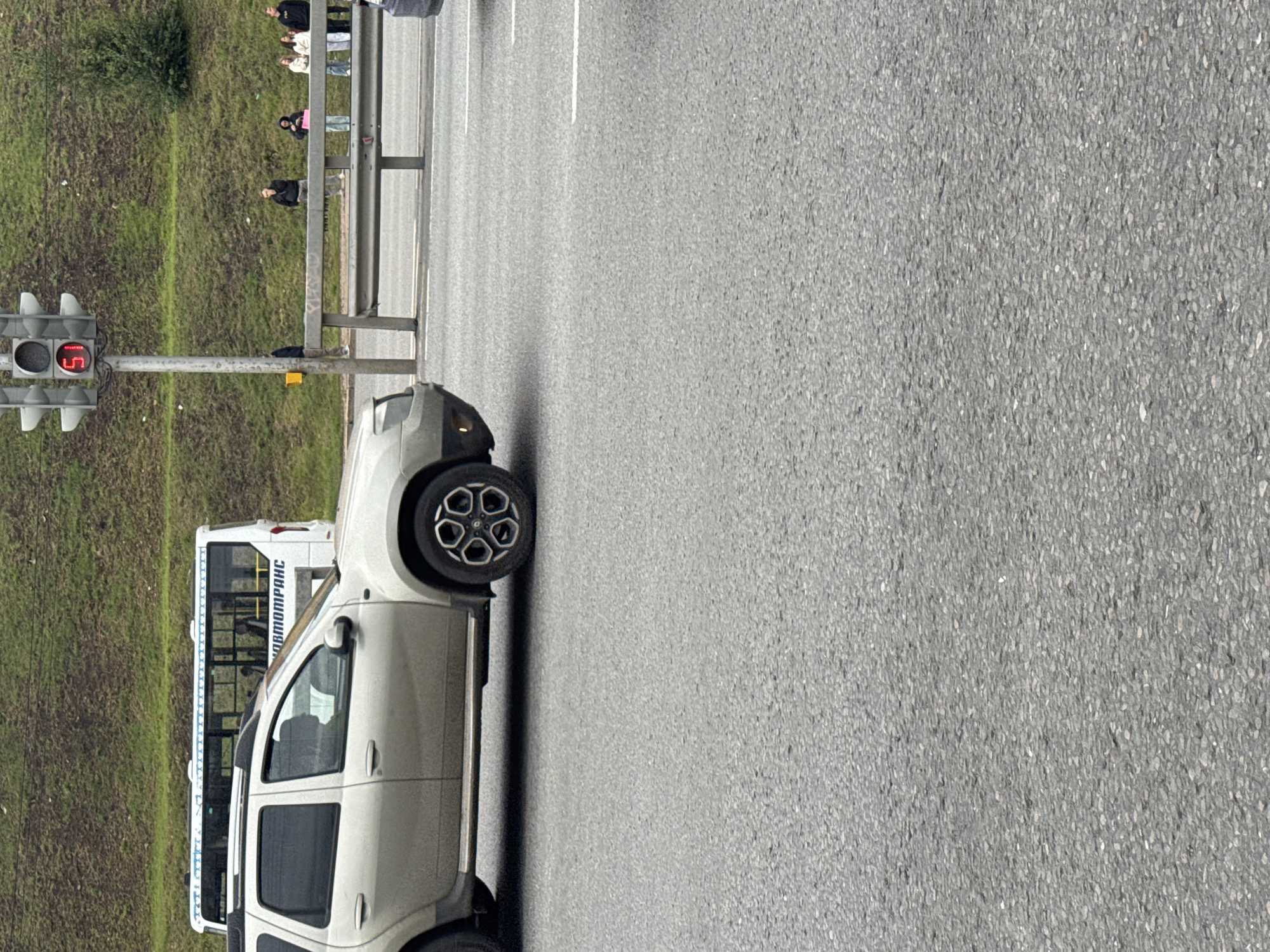 The image depicts a traffic scene on a multi-lane road, likely within the city of Ufa, Russia. A light-colored SUV is prominently featured in the foreground, driving on the asphalt road. Behind the SUV, a white bus with blue trim and the text "АВТОТРАНС" is visible. To the right of the bus, a traffic light displays a red signal with the number "51" illuminated. Several individuals are gathered near a guardrail on the grassy embankment behind the bus, suggesting a bus stop or waiting area. The environment is overcast, with a green, sloped embankment forming the background.