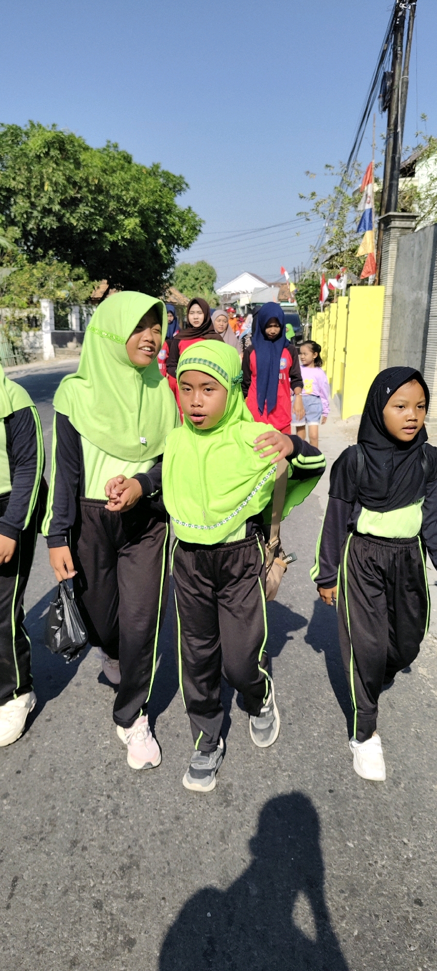 In Sulang, Indonesia, on a clear, sunny day, a group of children wearing matching black and green tracksuits and hijabs walk along a paved road. The children appear to be of elementary school age, and they are moving in the same direction. In the foreground, two girls in bright lime green hijabs and tracksuits are most prominent. The girl on the left, with her eyes open and mouth slightly agape, holds hands with the girl to her right. The second girl, also in a lime green hijab, looks forward with her mouth open, her right arm resting on the shoulder of the first girl. Behind them, another child in a black hijab and tracksuit is visible, her face turned to the right. Further back, a child in a red shirt and blue hijab is seen, along with other individuals, suggesting a larger group activity, possibly a school outing or religious procession. The environment is characterized by residential buildings with yellow walls and fences, trees lining the street, and utility poles with wires overhead. Indonesian flags are displayed on poles in the background. The road surface is asphalt, and shadows of the children are cast on it, indicating the time of day. The overall scene depicts a communal activity in a typical Indonesian neighborhood.