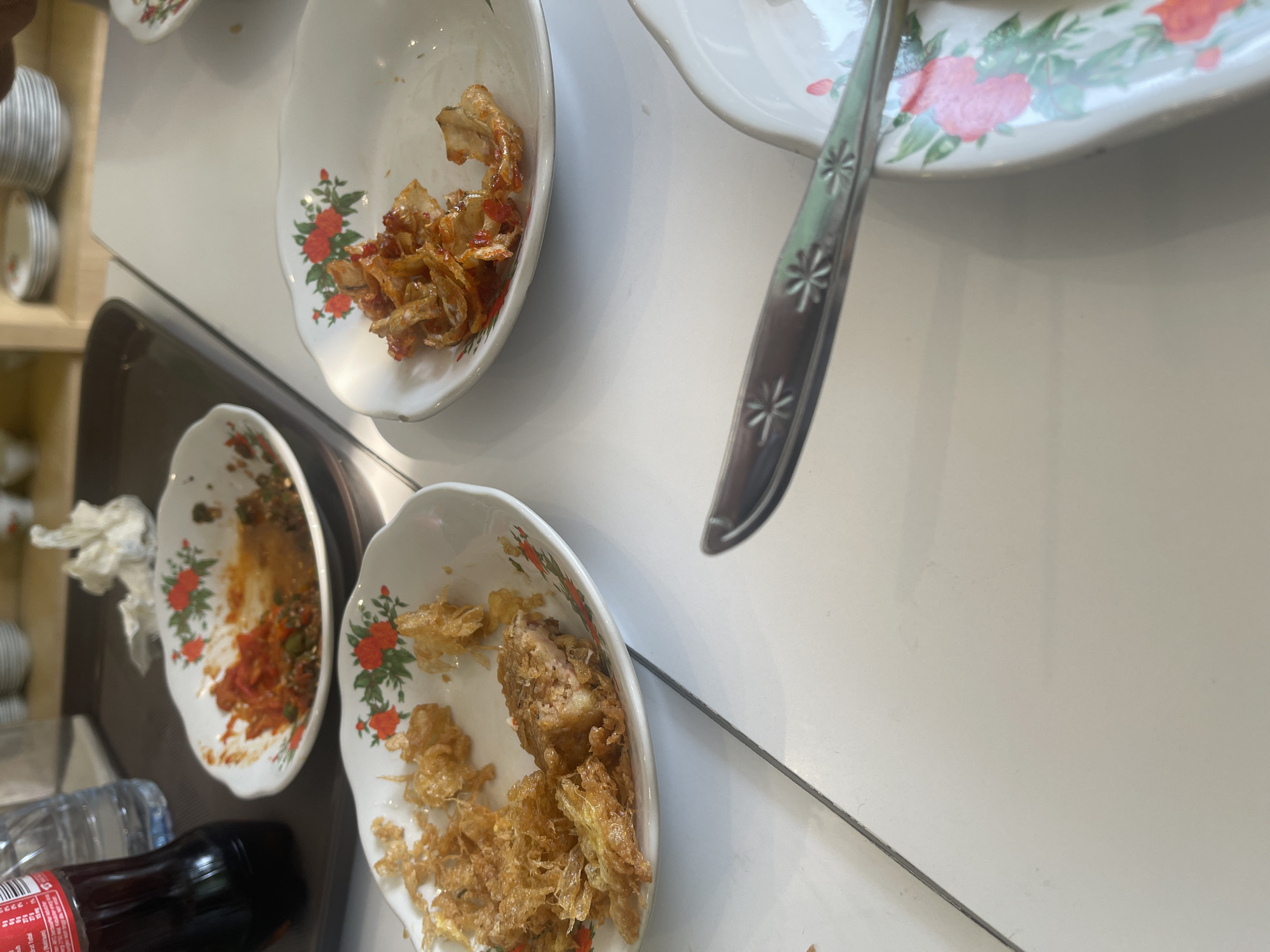 The image depicts a table with leftover food in several white, floral-patterned plates. In the foreground, a fork rests on the table, its handle showing a starburst pattern. To the left, a partially eaten meal is visible in a plate, consisting of crispy fried items and a piece of meat. Another plate to the right contains remnants of what appears to be shredded meat in a sauce. A third plate in the background shows traces of red sauce and green chili. A dark bottle of soda and a bottle of water are also present on a brown tray. The setting appears to be an indoor dining area, possibly a restaurant in Medan, Indonesia, given the context of the request. The overall scene suggests a meal has recently concluded, with remnants of food and drinks remaining on the table.