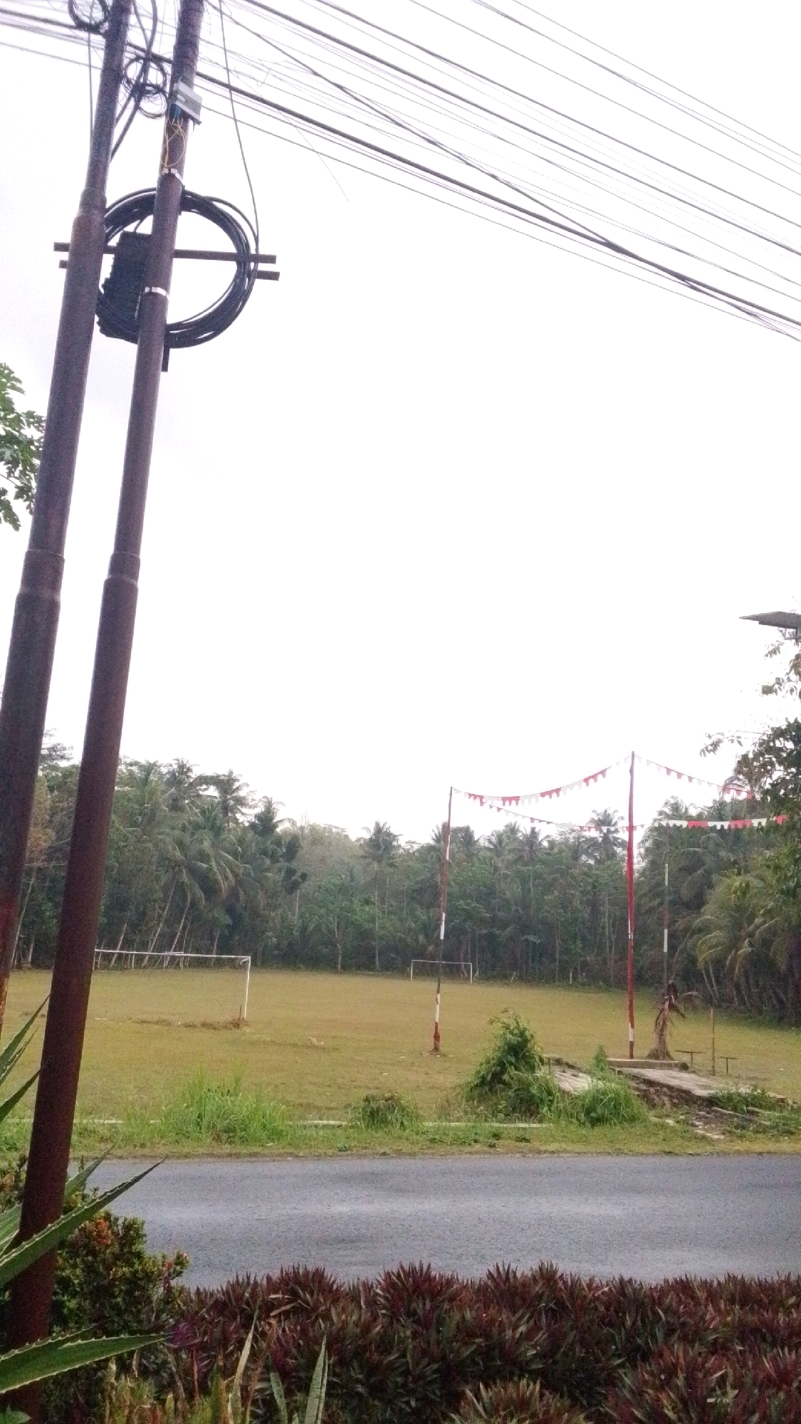 The image depicts an outdoor scene at what appears to be a field in Cintaratu, Indonesia. In the foreground, two large, reddish-brown utility poles stand vertically, adorned with coiled black cables and other electrical equipment. Overhead, a dense network of power lines stretches across the frame against a bright, overcast sky. Beyond the utility poles, a grassy field is visible, with two white soccer goal frames positioned at opposite ends. The field is bordered by a dense line of palm trees and other lush vegetation. In the middle ground, two tall poles are erected, draped with alternating red and white triangular flags, suggesting a festive or commemorative event. A paved road runs horizontally across the lower portion of the image, with a strip of dark, leafy plants in the immediate foreground. The overall atmosphere is subdued, likely due to the overcast weather conditions.