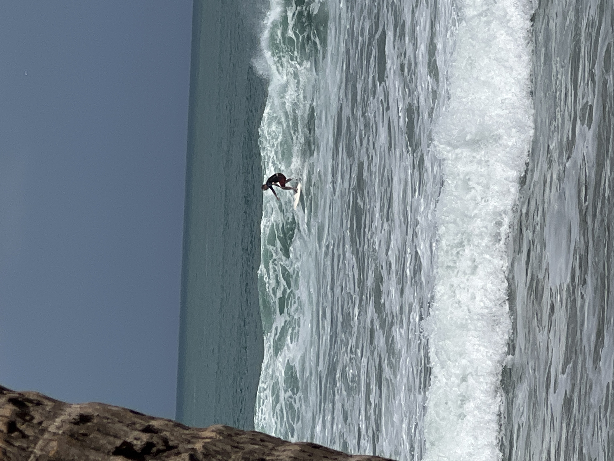 A lone surfer rides a wave in the ocean off the coast of Kuta, Indonesia. The surfer, wearing a dark top and red shorts, stands on a white surfboard. Waves are breaking around the surfer, with white foam scattered across the water's surface. In the foreground, a large wave is crashing, and the ocean extends to the horizon under a hazy blue sky. The edge of a large rock or piece of driftwood is visible in the lower left corner of the frame.