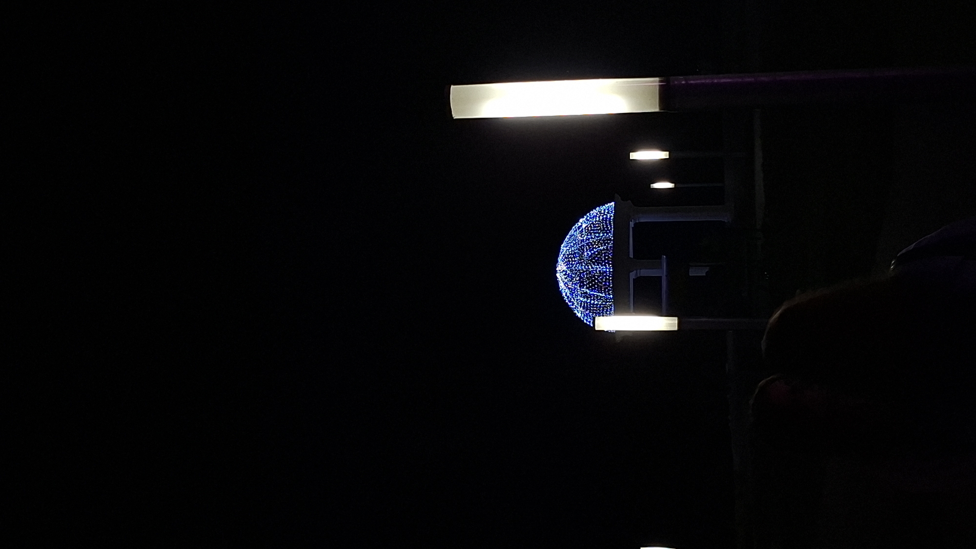 In Myadzyel, Belarus, a dark night scene features a gazebo adorned with blue lights. The gazebo is situated in the background, with its domed roof intricately covered in twinkling blue lights, creating a festive atmosphere. In the foreground and mid-ground, several tall, rectangular light fixtures with white illumination are positioned, casting a glow on the surroundings. The lighting creates a stark contrast against the dark night sky. In the lower portion of the image, two figures are partially visible, silhouetted against the ambient light. Their presence suggests a public or recreational area, possibly a park or a town square, illuminated for evening enjoyment. The overall impression is one of a nighttime gathering or event, with decorative lighting enhancing the urban landscape.