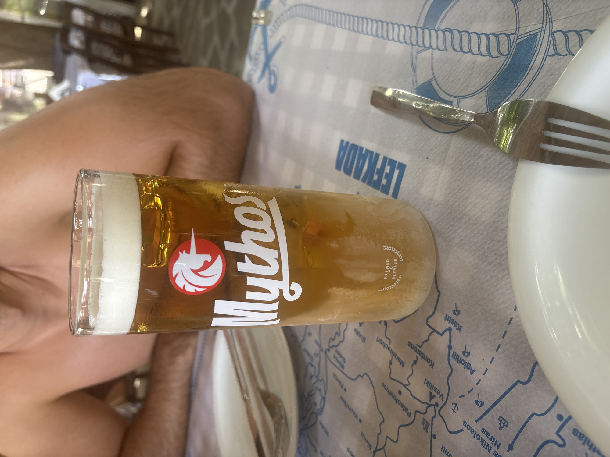 A person's bare shoulders and upper chest are visible in the background of a close-up shot focused on a glass of beer. The glass, filled with a golden-amber liquid topped with a thick layer of white foam, has the "Mythos" logo printed on it in white lettering, accompanied by a red circular emblem featuring a white griffin. Below the logo, the words "BREWED TO REFRESH" are visible. The glass sits on a tablecloth printed with a blue map of what appears to be Lefkada, Greece, highlighting various locations such as "Paleofoumos," "Marathocchori," and "Kastri." A fork rests on the right side of the table, partially obscuring a design on the tablecloth that includes a life preserver. In the background, a white plate and silverware are also visible. The scene suggests an outdoor dining setting.