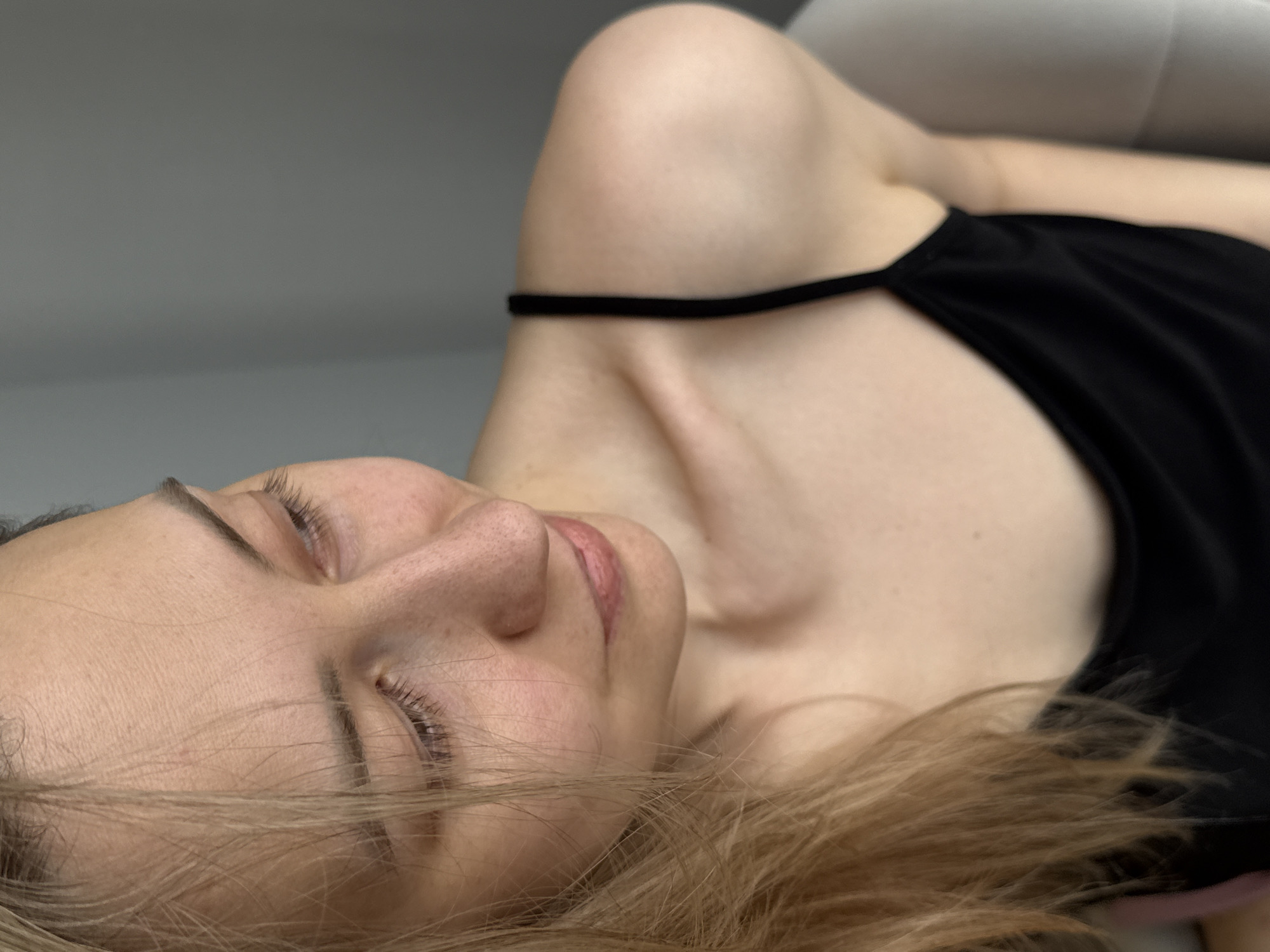 A woman lies on her back. Her eyes are closed. She has fair skin and blonde hair. She is wearing a black tank top. The background is a light gray wall. The location cannot be determined from the image.