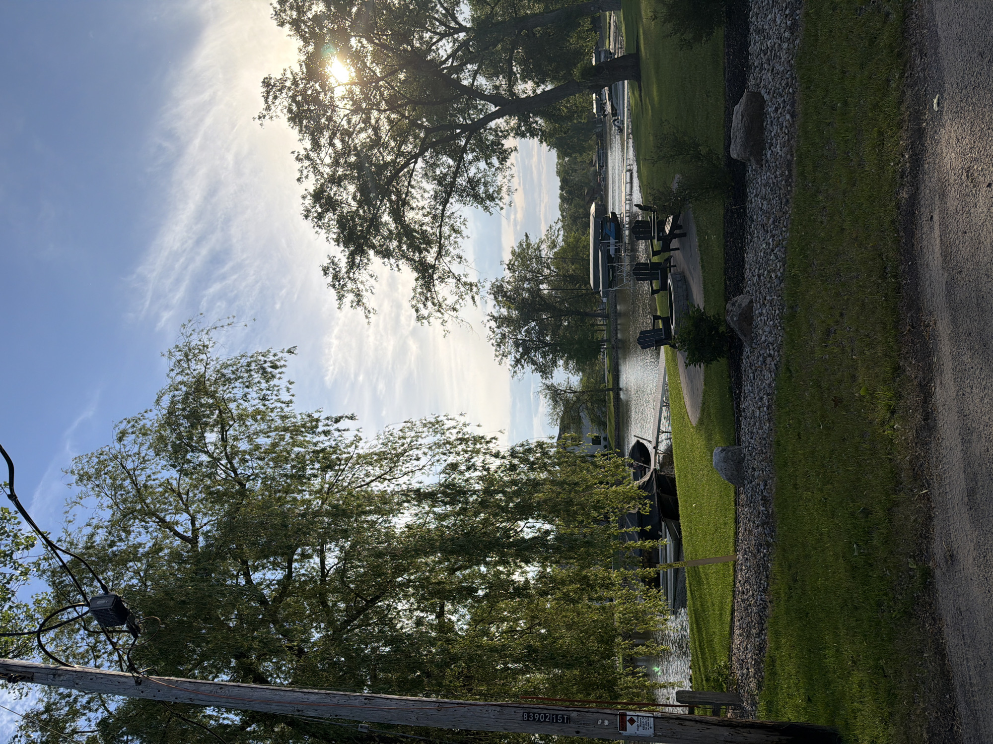 This photograph captures a serene, sun-drenched lakeside or canalside scene. The image is composed in a landscape orientation (after correcting the original rotation), with a clear blue sky and wispy white clouds dominating the upper portion. The sun is visible in the upper right, partially obscured by the lush green foliage of a large tree, creating bright sunbeams and a warm glow.
In the midground, a calm body of water stretches horizontally, reflecting the sky and surrounding greenery. Several boats, some covered, are moored along the grassy bank, indicating a residential or recreational area. Adjacent to the water, on a well-maintained lawn, are a few black Adirondack chairs and what appears to be a matching fire pit, suggesting an outdoor seating area. Decorative rocks are scattered along the edge of the grassy area, bordering a dirt or gravel path on the right side of the frame.
In the foreground on the left, a wooden utility pole with electrical wires and a small black equipment box is visible. Text "B39021ST" is clearly legible on the pole. Below this, a red-bordered warning sign, featuring a white background and a black symbol that appears to be a hand touching electrical current, is partially visible.
No people are present, lending a peaceful and tranquil atmosphere to the scene. The bright sunlight suggests it is either morning or late afternoon. The location cues, such as the boats, docks, and utility pole, point to a developed area alongside a natural water body in Lake Delton, United States of America.