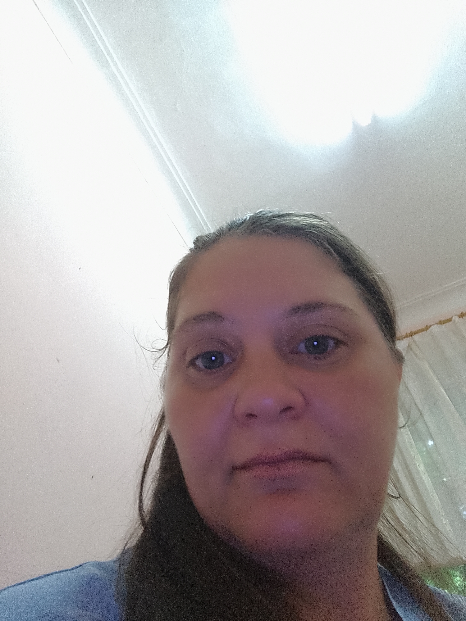 A close-up, low-angle, indoor selfie shows a woman from the chest up. She has brown hair pulled back, exposing her face, which is tilted upwards. The woman is wearing a blue shirt. The background is a white wall with a crown molding at the top, and a sheer curtain is visible on the right side of the image. A bright light source, likely overhead, is causing some overexposure in the upper portion of the image.