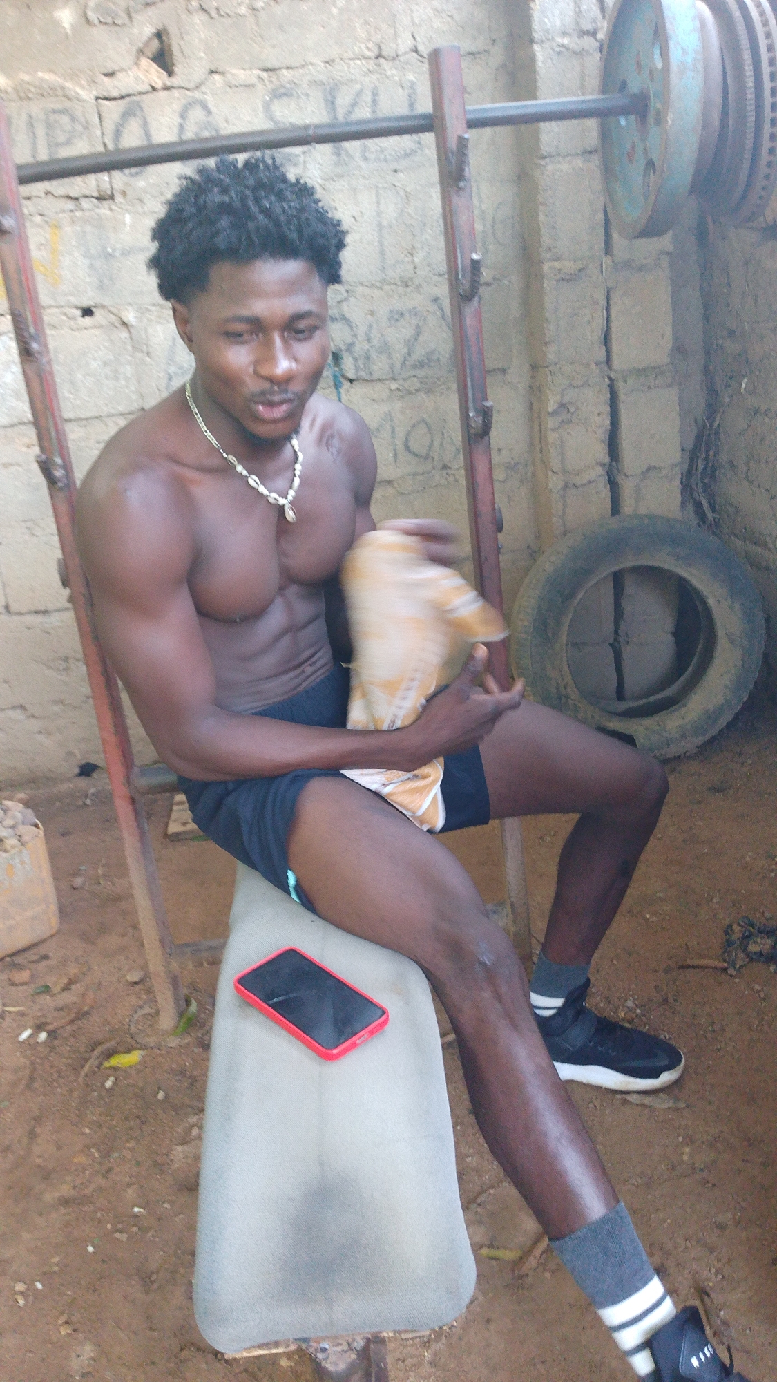In Akwanga, Nigeria, a young man with dark, curly hair and a muscular physique is seated on a cushioned bench. He is shirtless, wearing dark shorts, grey socks with white stripes, and black athletic shoes. Around his neck, he has a necklace with a shell pendant. In his right hand, he holds a beige and orange cloth. To his left, a red-cased smartphone lies on the bench.
The background suggests an outdoor or semi-outdoor gym setting. A metal weightlifting rack is visible, with weight plates mounted on the left side. A discarded tire rests on the ground behind him. The wall behind the rack is constructed of rough-hewn bricks or concrete blocks, with some scribbled markings on its surface. The ground is composed of packed earth and sand, with some small stones and debris scattered around. The overall environment appears rustic and functional, likely a local gym or workout area.