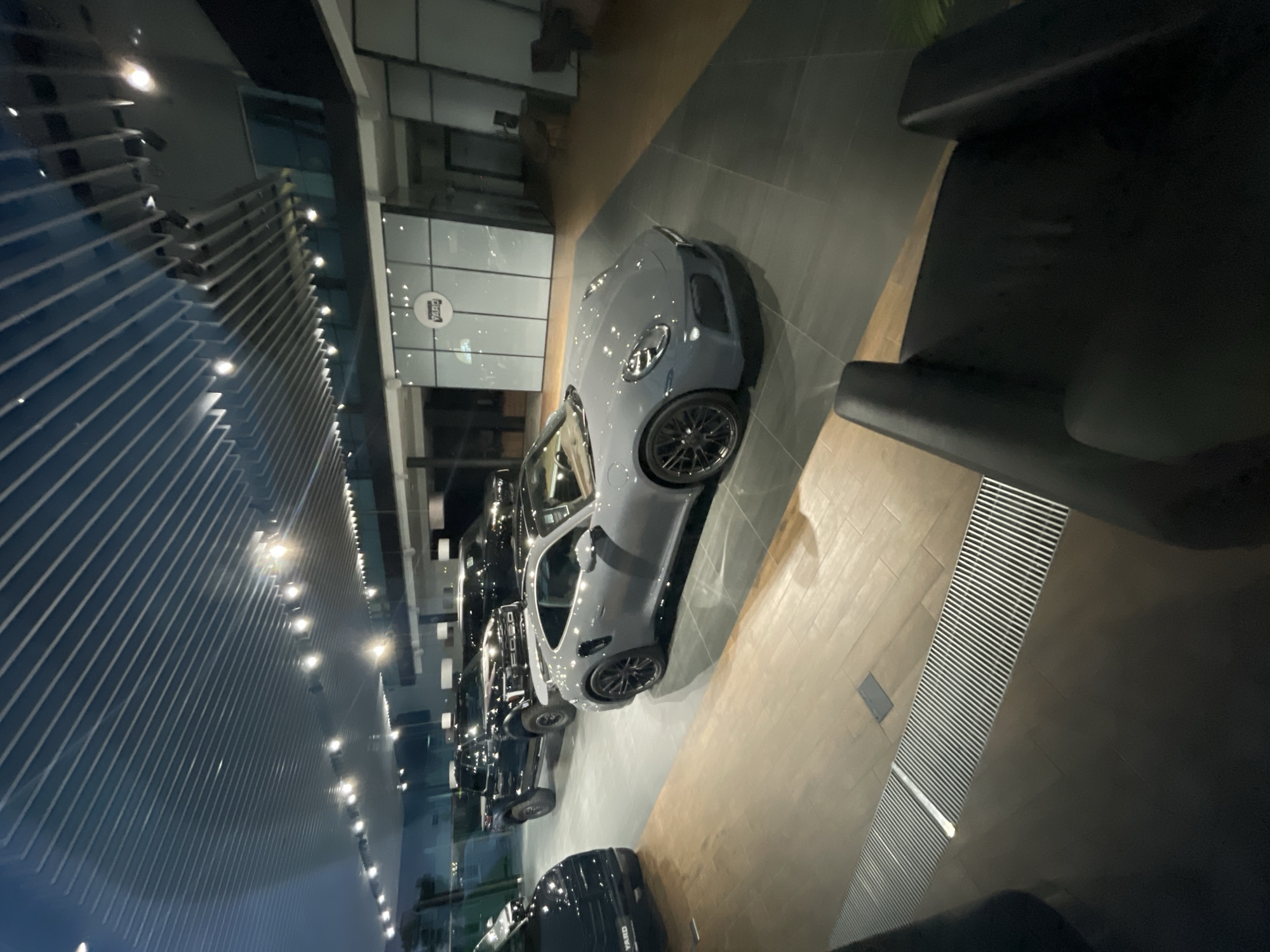 This is a medium shot, eye-level, interior photograph of a modern car dealership showroom. The floor is a combination of polished wood planks and dark grey tiles. The ceiling features a contemporary design with parallel, angled metal slats and recessed lighting. In the foreground, a dark grey luxury sports car, possibly a Porsche, is parked on the tiled floor, facing towards the left of the frame. Its headlights are illuminated. Behind it, a black pickup truck, identified by "FORD" on its grille, is parked. Further back, other vehicles and a section of the dealership's interior, including glass partitions and a logo that appears to read "YARD," are visible. The overall atmosphere is clean, well-lit, and upscale, typical of a high-end automotive retail space. There are no people visible in the image.