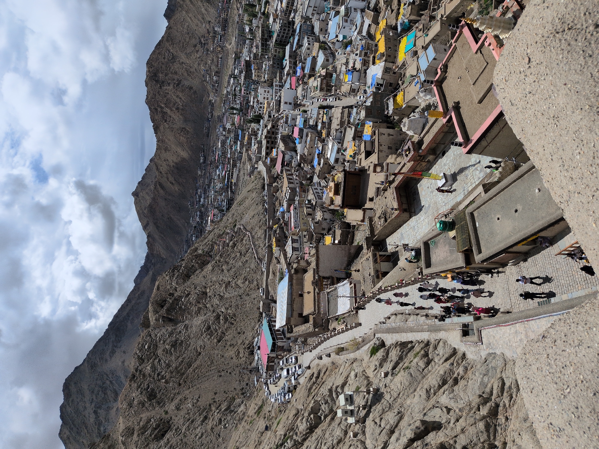 The image depicts a high-angle view of Leh, India, with a dense collection of buildings nestled at the base of rugged, barren mountains under a cloudy sky. In the foreground, a stone wall or platform is visible, with a walkway leading down to a street lined with parked cars and buildings. People are seen walking along the path and in the street. The architecture of the buildings is a mix of traditional and modern, with flat roofs, some adorned with colorful tarps (yellow and blue). The street is bustling with activity, indicated by the presence of numerous vehicles and pedestrians. Further into the cityscape, more buildings stretch across the valley, climbing the slopes of the surrounding mountains. The environment is dry and arid, characteristic of the Himalayan region. The overall scene conveys a sense of a vibrant, albeit densely populated, mountain town.