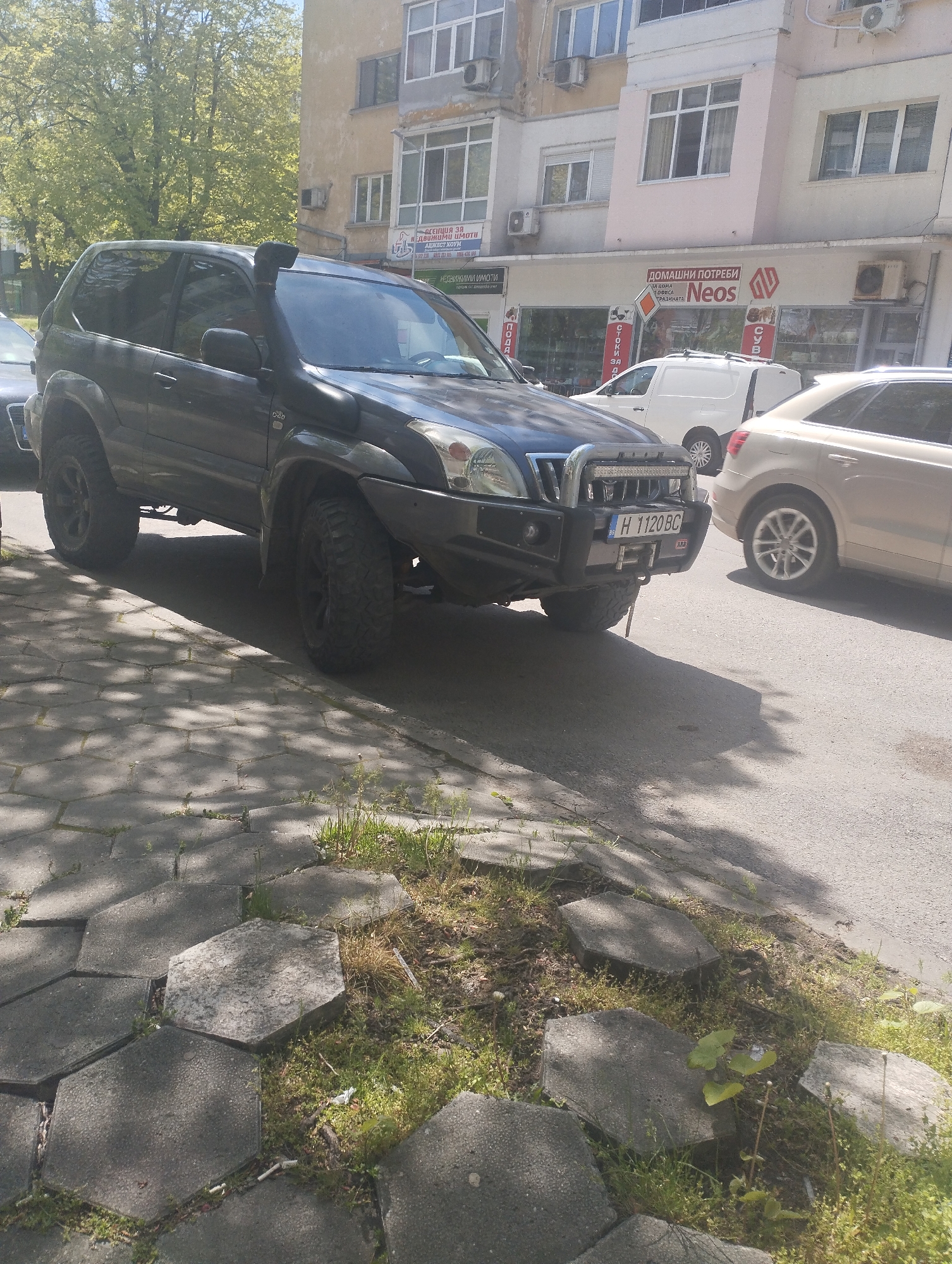 The image captures a street scene in Shumen, Bulgaria, where a heavily modified dark-colored Toyota Land Cruiser Prado is parked on the sidewalk. The vehicle features off-road modifications, including a snorkel, upgraded bumper with a winch, and off-road tires.
Behind the Toyota, there's a beige Audi SUV and a white van, both parked on the street. A building lines the street in the background, with storefronts visible on the ground floor. One store has "Домашни Потреби NEOS" written on it, which translates to "Household Goods NEOS." Another business appears to deal with real estate, indicated by the sign "Недвижими Имоти" ("Real Estate").
The sidewalk is paved with hexagonal stones, some of which are surrounded by grass and weeds. The lighting suggests it's a sunny day. The license plate of the Toyota reads "H 1120 BC."