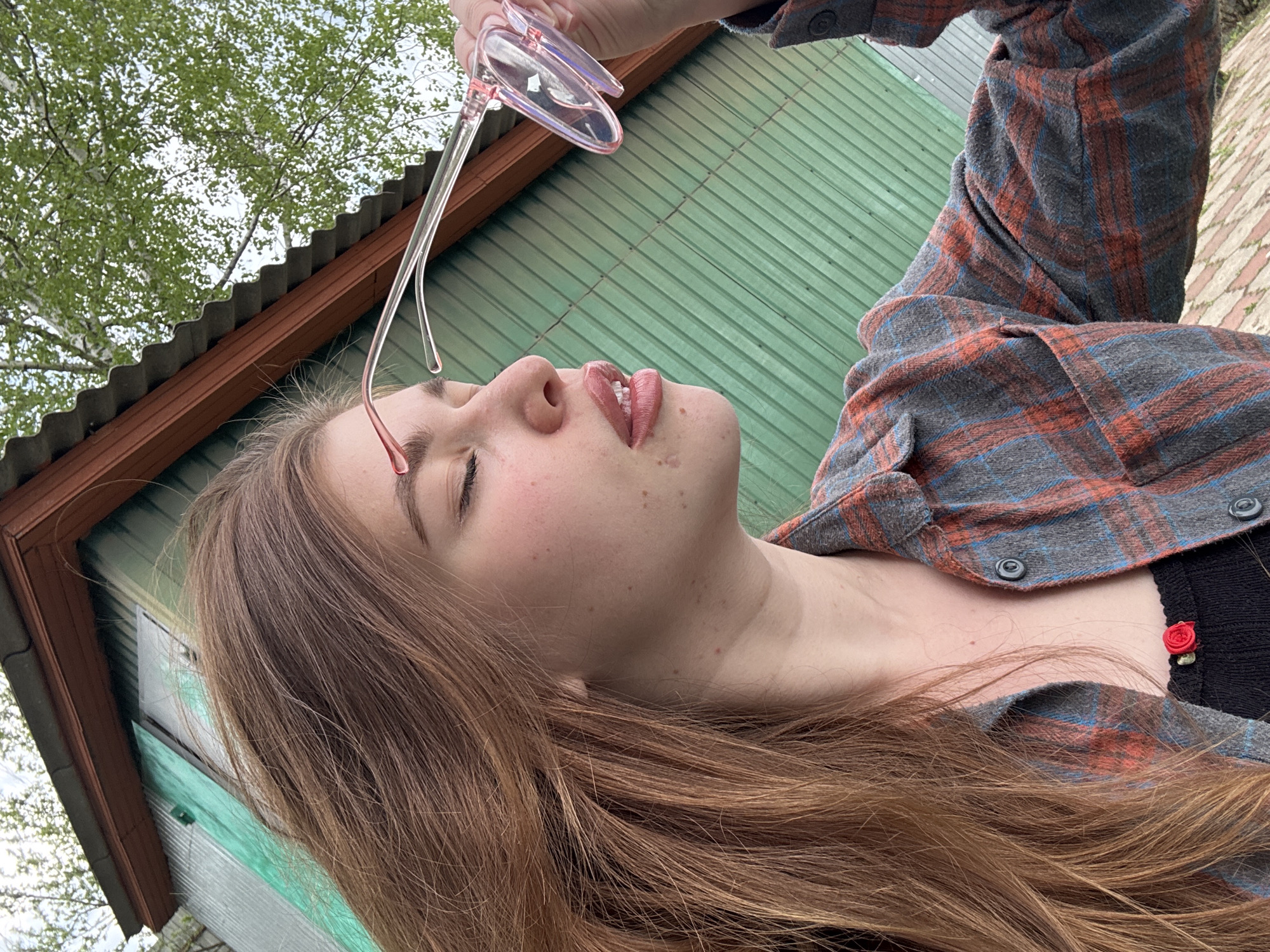 A young woman with long, light brown hair is outdoors, holding a pair of pink-framed eyeglasses above her face. Her eyes are closed, and her head is tilted back. She is wearing a plaid shirt over a black top with a small red flower detail. The background shows a corrugated metal wall, a roof overhang, and a tree with green leaves. The location appears to be in a rural setting. The image provides no information allowing a determination of whether this scene is located in the city of Мещёры, Russia.
