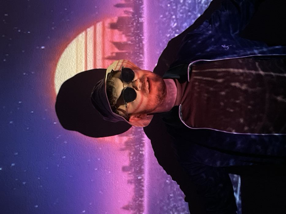 A man wearing a bucket hat and sunglasses is positioned against a backdrop reminiscent of a retro-futuristic cityscape. The scene features a large moon and a skyline rendered in vibrant pinks and purples. Stars dot the sky above the buildings. The man's jacket is dark with a light-colored trim. The lighting projects the cityscape pattern onto his shirt.