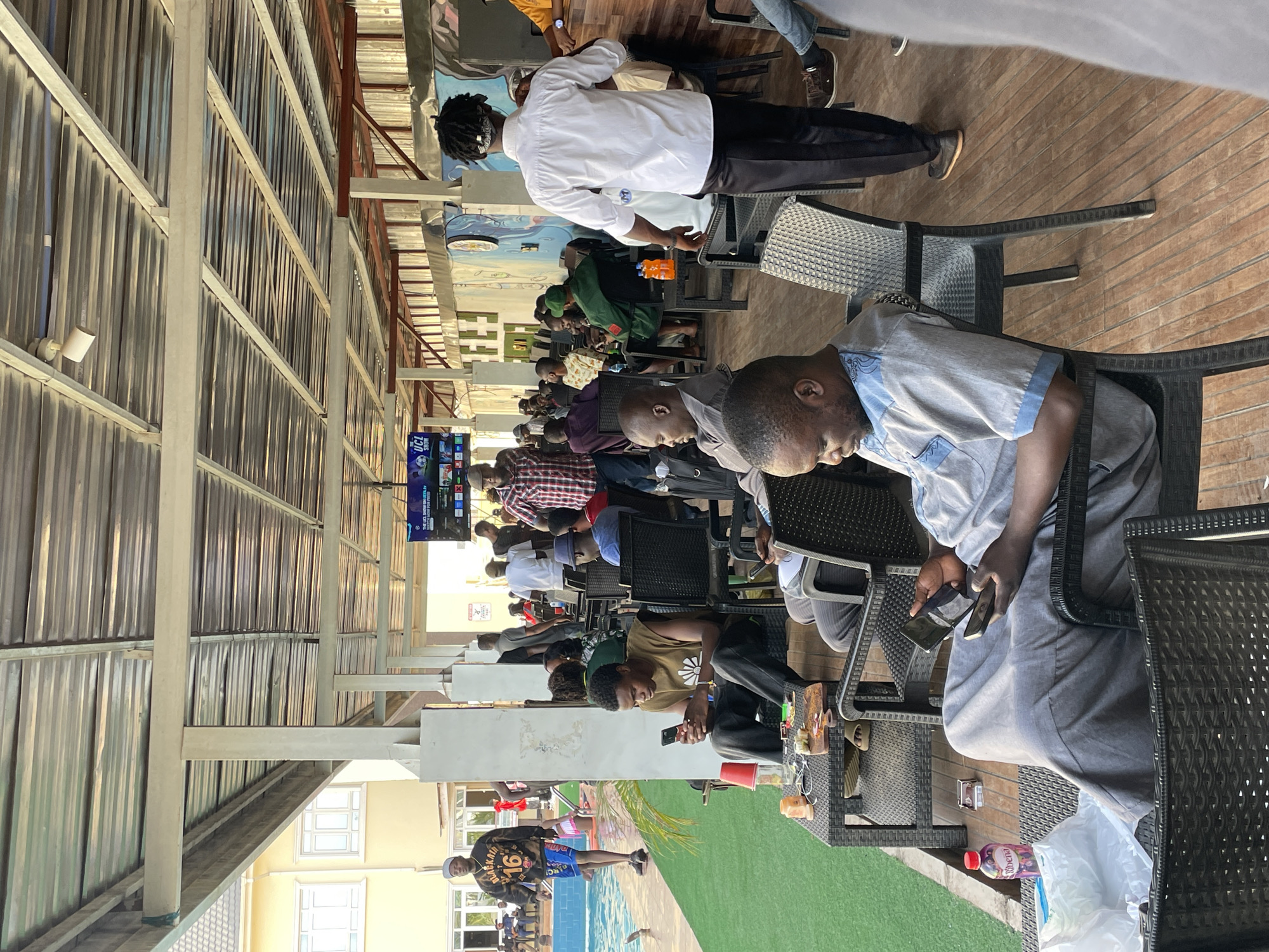 The image shows an outdoor seating area in Bar Jirgi Summa, Nigeria. Multiple people are seated at tables, many appearing to be looking at or interacting with their mobile phones. A television screen is visible in the background, displaying sports content. There are chairs and tables made of woven material. A pool is visible outside the covered area, and at least two people are standing near the pool. The area is covered with a metal roof, supported by pillars. There are drinks and personal belongings on the tables.