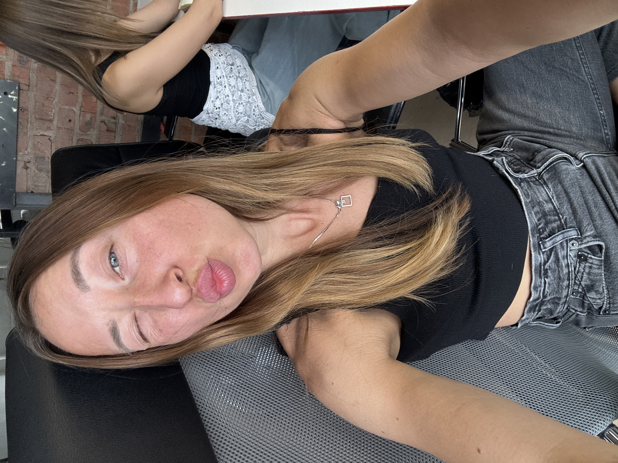 A woman is sitting in a chair, facing the camera with a sideways angle, winking and puckering her lips. She is wearing a black top and gray jeans. Her hair is light brown. Part of another woman is visible behind her, wearing a black top and white patterned garment, sitting at a white table. A brick wall is visible in the background. The location is Chaikovsky, Russia.