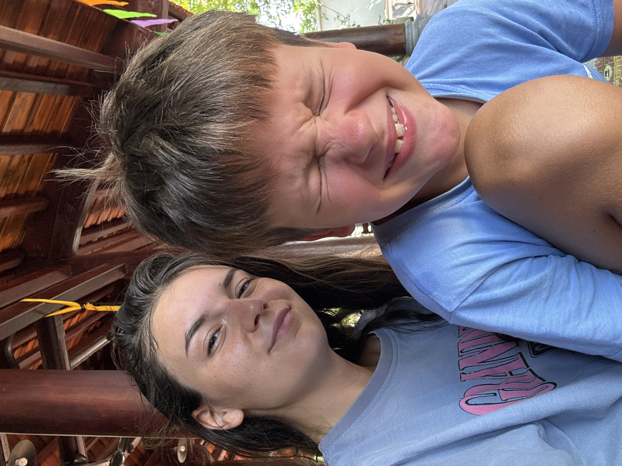 The image depicts two individuals, a young boy and a woman, likely posing for a selfie in Mũi Né, Vietnam. The boy, with short brown hair, is positioned at the top of the frame and is squinting with a broad smile. He is wearing a light blue t-shirt. The woman, who is at the bottom of the frame, has long brown hair, fair skin, and a neutral expression. She is wearing a blue t-shirt. The background indicates a location with wooden architectural elements, suggesting an outdoor or semi-outdoor setting. Multi-colored flags are visible in the upper left corner, hinting at a festive atmosphere. The overall composition appears casual and spontaneous.