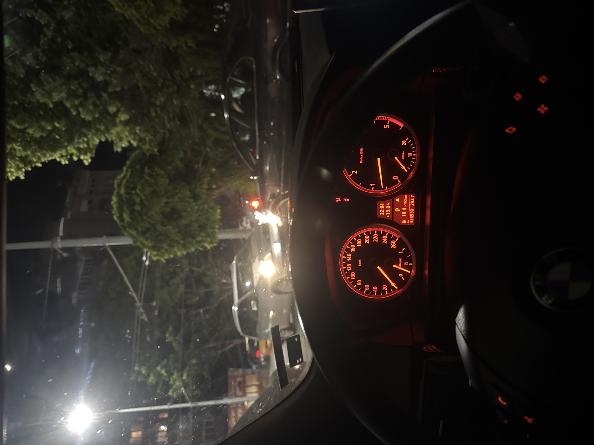 The image shows the interior of a BMW car, specifically the dashboard and gauges, at night. The dashboard displays show the car is in Park, the engine is idling around 0 RPM, and the car's speed is 0 km/h. The temperature outside is 19 degrees Celsius, and the car has traveled 328,930 kilometers.
Outside the car, through the windshield, there are streetlights, trees, and other vehicles. The environment suggests an urban setting. The presence of other vehicles and trees indicates a possible street scene in Stara Zagora, Bulgaria. The image likely captures a moment while the car is stationary at night, potentially in traffic or parked.
