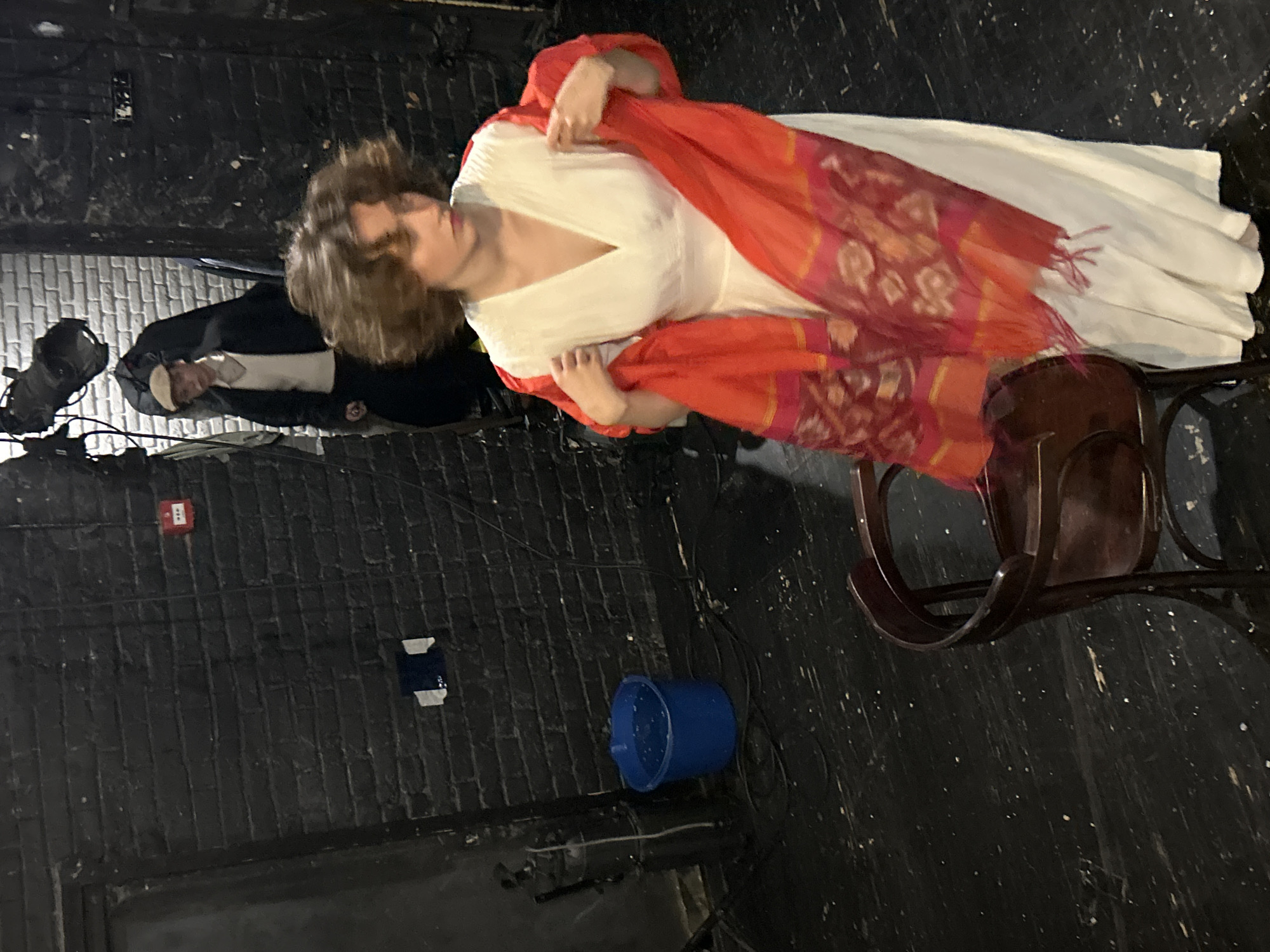 In a dimly lit indoor space with black brick walls and a dark wooden floor, a woman in a white dress is holding a vibrant red and orange scarf. Her hair is dark and curly, and she is looking downwards to her right. To her left, an empty dark wooden chair is positioned. In the background, a person wearing a light-colored sweater, dark jacket, and a flat cap is walking away from the camera. There is a blue bucket filled with water on the floor to the left, and theatrical lighting equipment is visible hanging from the ceiling in the upper part of the image. A red fire alarm is mounted on the brick wall. The overall environment suggests a backstage area or a studio setting.