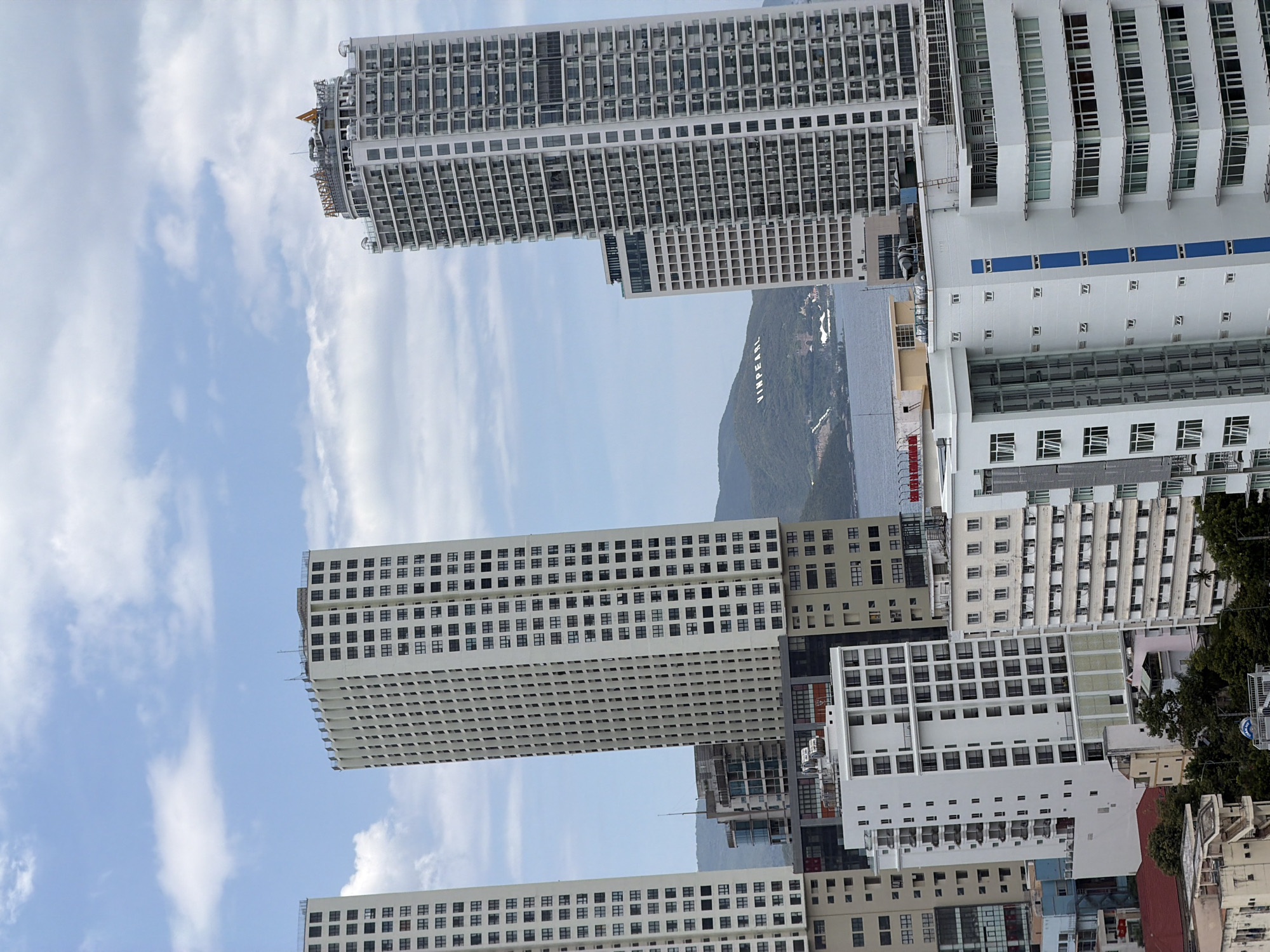 This is an aerial shot of a coastal city skyline in Nha Trang, Vietnam. In the foreground, multiple modern high-rise buildings dominate the scene, varying in height and architectural style. Several of these are residential or hotel towers with numerous balconies and windows. One prominent building on the right displays the name "VINPEARL" in large white letters on a lush green hillside in the background. A body of water, likely the ocean, is visible beyond the buildings and in front of the hillside. The sky is a bright blue with scattered white clouds. The overall impression is one of urban development and coastal tourism.