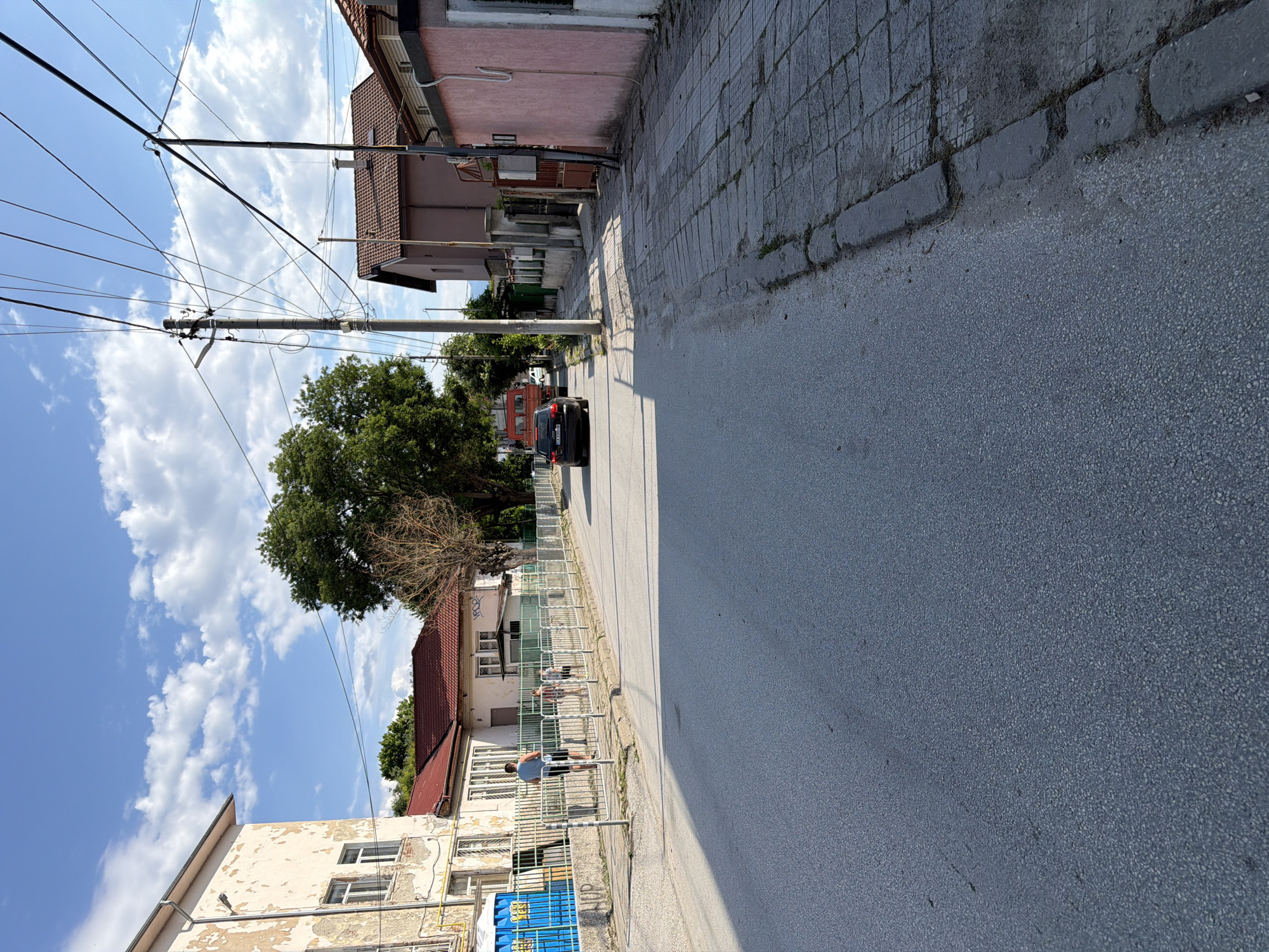 The image depicts a residential street in Plovdiv, Bulgaria, under a clear blue sky with scattered white clouds. A paved road runs horizontally through the center of the frame, bordered by sidewalks on both sides. On the left, an older, light-colored building with peeling paint and multiple windows is visible, along with a blue portable toilet behind a metal fence. A few people, including a man in a blue t-shirt and black shorts, and two children, are present near the fence. A green metal fence runs along the sidewalk.
The right side of the street features a pink building with a dark metal gate and a brown tiled roof. Further down the street, a light brown building with a similar tiled roof is visible. A large tree with dense green foliage stands between the buildings.
Several utility poles with a complex network of overhead wires are prominent features of the scene. Two vehicles are on the road: a black car in the foreground and a red truck further in the distance. The sidewalk on the right is paved with large, irregular stone slabs, some of which appear worn and uneven. The overall impression is of a quiet, lived-in urban environment.