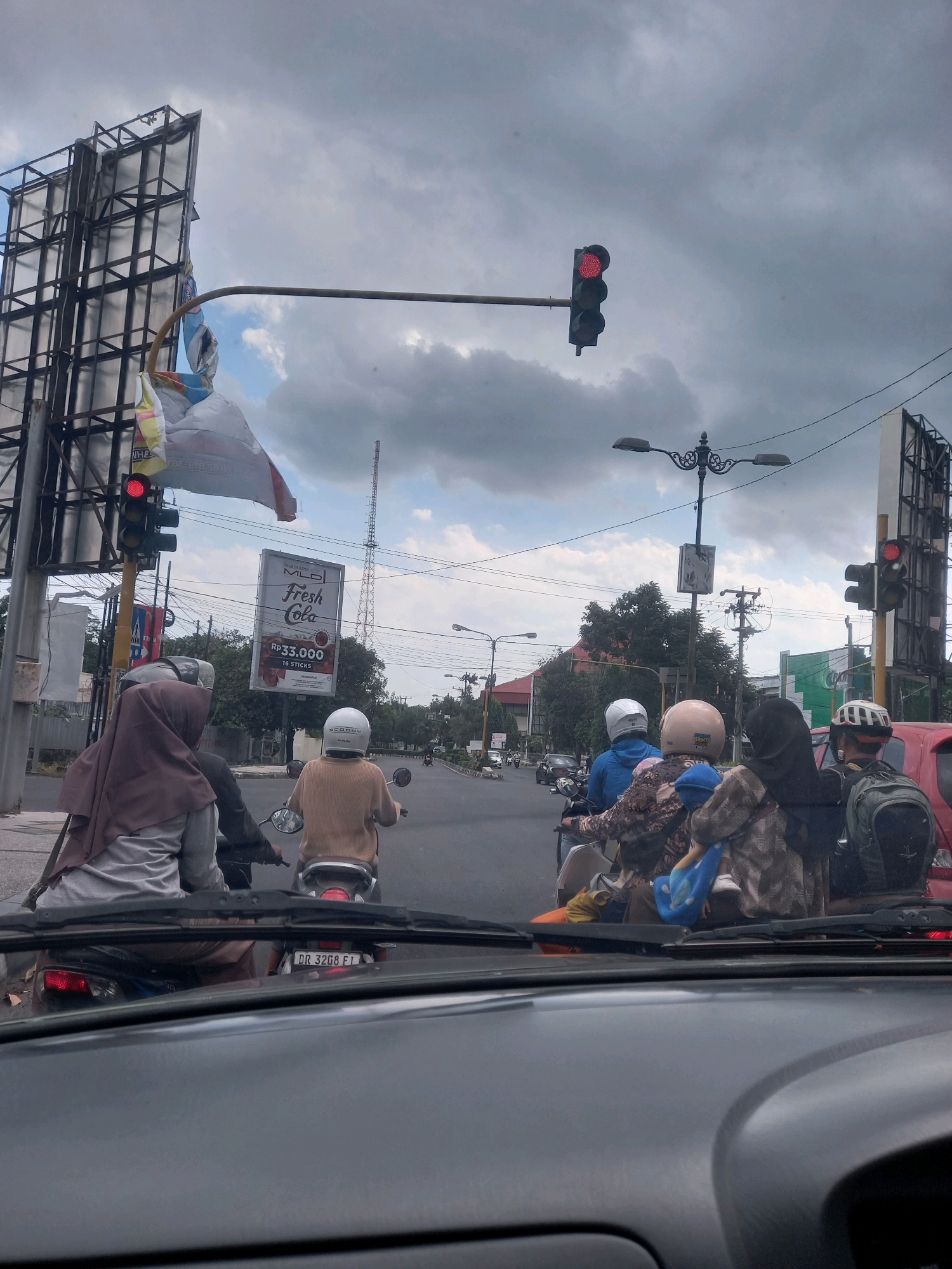 The image shows a street scene in the city of Mataram, Indonesia, on a cloudy day. Several motorcycles are stopped at a traffic light, which is red. The perspective is from inside a vehicle looking forward.
In the foreground, the dashboard of a car is visible. On the road, multiple motorcycles are waiting at the intersection. The first motorcycle on the left has two passengers, one wearing a brown hijab. The second motorcycle has a driver wearing a light brown shirt and a white helmet, and a passenger behind them. Further ahead, another motorcycle carries a driver in a blue jacket and two passengers, one wearing a patterned jacket and the other a black hijab. To the right, a red car is also stopped.
Various elements of urban infrastructure are present: a large billboard structure stands to the left, partially covered by a torn advertisement banner. A traffic light is clearly visible, displaying a red signal. Streetlights and utility poles with tangled wires are also scattered throughout the scene. In the background, trees and buildings are visible, along with a telecommunications tower. A billboard for "MLD Fresh Cola" priced at Rp 33.000 for 16 sticks is also displayed. The license plate of the motorcycle in front of the viewer's vehicle reads "DR 32G8 ET". The overall atmosphere is that of a typical busy intersection in an Indonesian city.