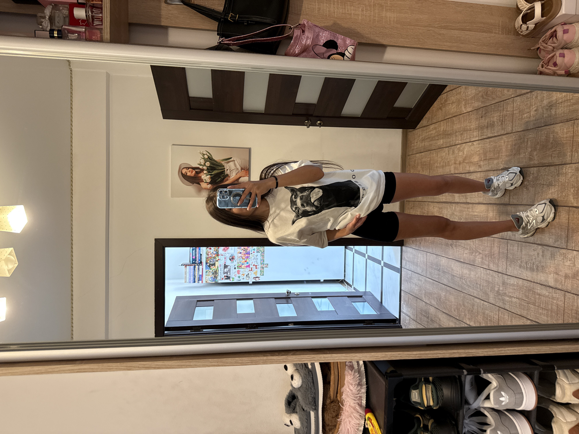 A young woman is taking a mirror selfie in what appears to be a bedroom in Chișinău, Moldova. She is wearing a white t-shirt with a graphic of a dog and black shorts. She has long, dark hair and is holding a smartphone. To her left, a shoe rack is visible, holding several pairs of shoes and slippers. Further to the left, a doorway leads into another room, with a patterned floor. To her right, a closet door with vertical panels is partially open. On the wall to the right of the closet, a black handbag and a pink Minnie Mouse themed bag are hanging. A wooden shelf above the bags holds various perfume bottles. The overall environment suggests a residential interior with personal belongings arranged around the space.