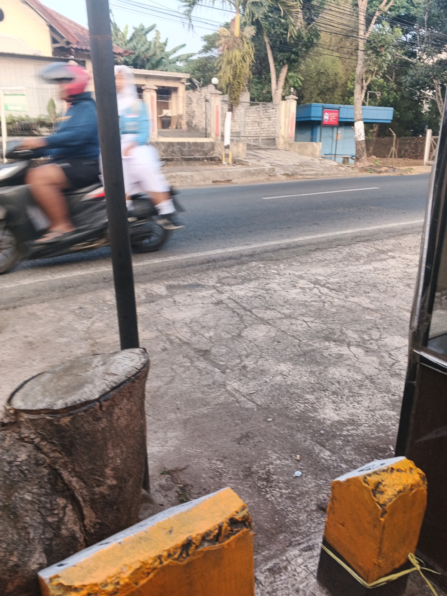 A motion-blurred, eye-level, outdoor shot shows a street scene in Semarang, Indonesia. In the foreground, on the left, is a tree stump. To its right, and further back, are two orange, concrete blocks with yellow string tied around them. A black pole, likely a traffic signpost, runs vertically down the center of the image.
In the mid-ground, a person wearing a blue jacket and red helmet rides a black motorcycle past a building with a white wall and a red-tiled roof. Behind the motorcycle rider, another person wearing a white hijab and a blue and white uniform is visible. A fence with stone pillars runs along the side of the street.
In the background, a blue building with a red sign is visible. Trees and foliage are scattered throughout the scene, indicating a relatively green urban environment. The road in the mid-ground has a white line marking.
