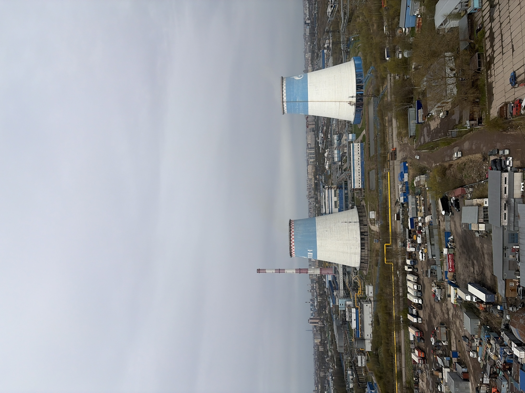 This image, taken from an elevated perspective, captures a sprawling industrial landscape, dominated by a thermal power plant under an overcast sky. The scene is set in what appears to be a city's industrial zone, with a distant city skyline visible on the horizon.
The most prominent features are two large, hyperboloid cooling towers, both predominantly white with a distinct blue band and white stripe near their tops. On the blue band of the lower cooling tower, the Cyrillic text "ТЭЦ-1" is clearly visible, identifying it as "Thermal Power Plant 1". A tall, slender smokestack, painted with alternating red and white horizontal stripes, stands between the two cooling towers.
Surrounding these central structures is a dense network of industrial buildings, warehouses, and storage facilities. Many buildings have blue or grey roofs. Various roads, some paved and others appearing to be dirt tracks, crisscross the area. Along these roads and in designated lots, numerous vehicles are parked, including a long line of white semi-trailer trucks, smaller cargo vans, and cars. Yellow pipes run along the ground in several sections, likely part of the plant's infrastructure. Scattered, bare or sparsely leaved trees and bushes indicate either early spring or late autumn.
The sky is a uniform, light grey, suggesting a heavily overcast day. No direct sunlight is visible, and shadows are soft or absent. There are no people visible, and no specific activities are actively taking place, giving the scene a static, observant quality. The overall impression is one of a large, functional industrial complex in Saint Petersburg, Russia, captured during daytime.