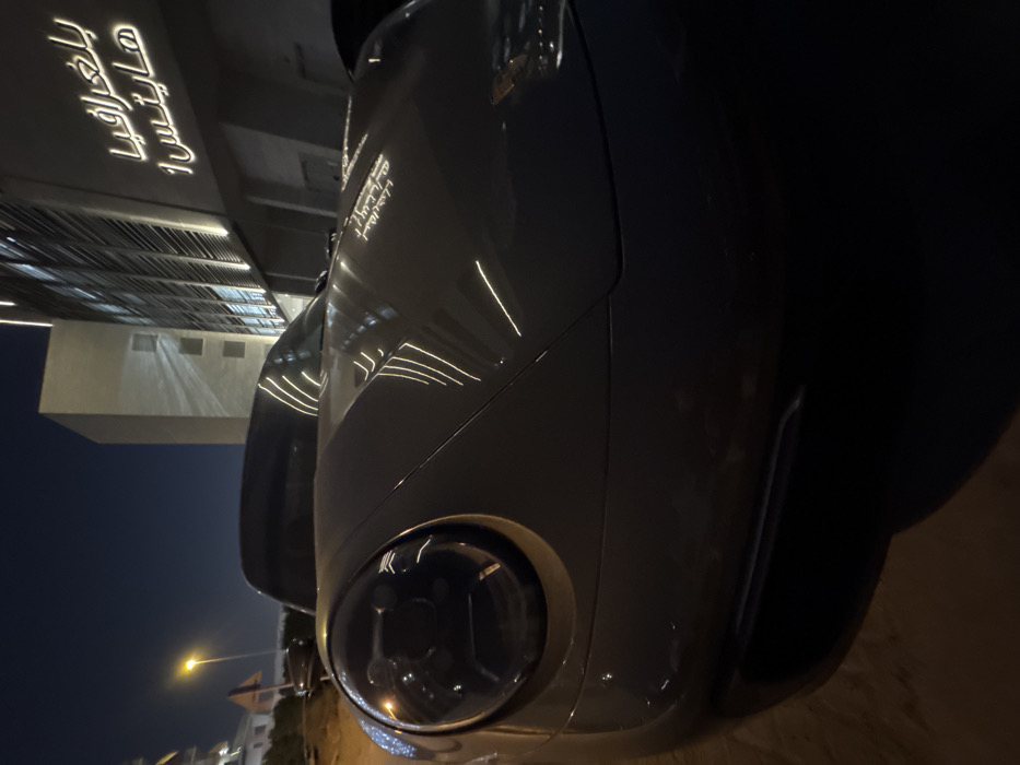 A close-up of a grey car at night with illuminated architectural reflections on its body. The round headlight dominates the foreground, while in the background, a building features horizontal light lines and Arabic script. A street lamp is visible in the distance, highlighting a section of the road. The image is oriented vertically.