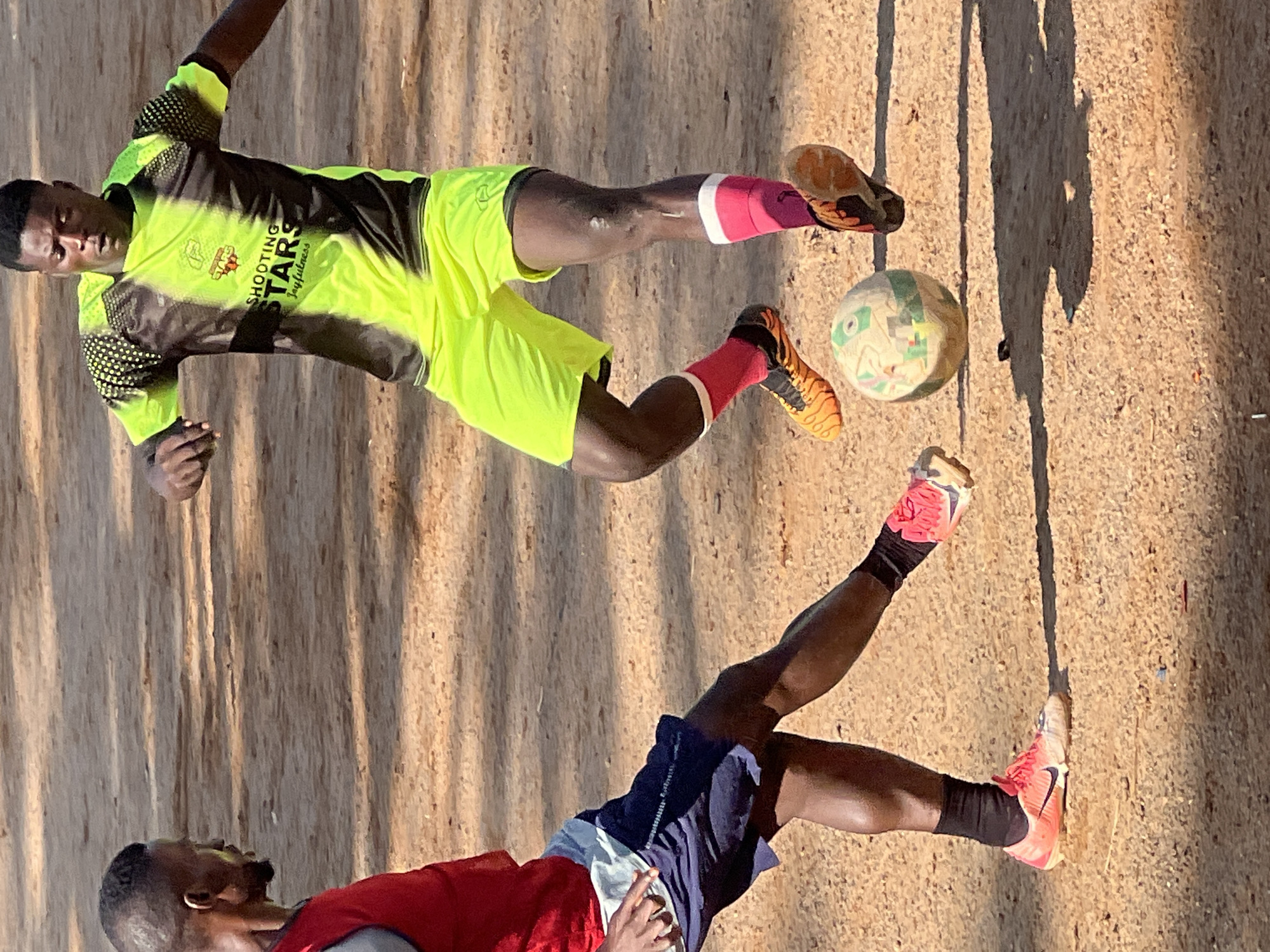 Two male individuals are engaged in a football match on a dirt field in Jos, Nigeria. The individual on the left wears a red vest over a grey shirt, navy blue shorts, and pink cleats. The individual on the right wears a neon yellow and black patterned jersey with "SHOOTING STARS" and "Joyfulness" printed on it, neon yellow shorts, and orange and black cleats with pink socks. A football lies on the ground between them, with the right player's foot positioned to kick it. The ground is covered in coarse, sandy dirt. Sunlight casts long shadows across the field.