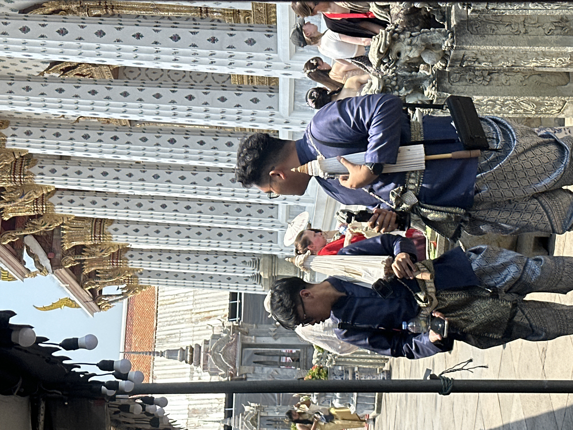 A man wearing a navy blue traditional Thai outfit, including a patterned sash and pants, stands holding a camera and a white umbrella. He is one of two men similarly dressed, standing on a paved area. In the background are ornate white and gold architectural structures with decorative patterns, characteristic of Thai temples. Further in the background, other people and temple details are visible. The photograph appears to be taken at Wat Arun, a Buddhist temple in Bangkok, Thailand, known for its intricate design and riverside location.