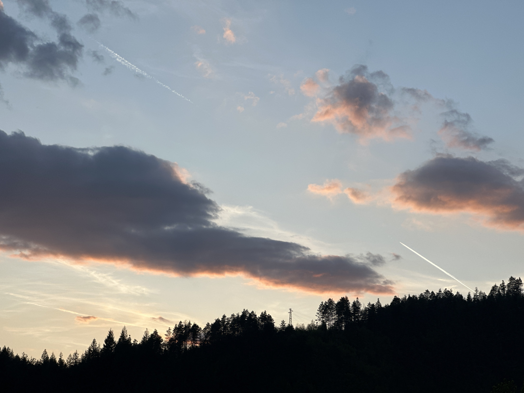 The image depicts a landscape, likely at sunset or sunrise, with a dark silhouetted treeline against a sky filled with clouds tinged with hues of orange and pink. Two contrails from airplanes are visible against the light blue sky. A power line pylon can be seen on the horizon. It's difficult to confirm the location specifically as Rudozem, Bulgaria without more context within the image.