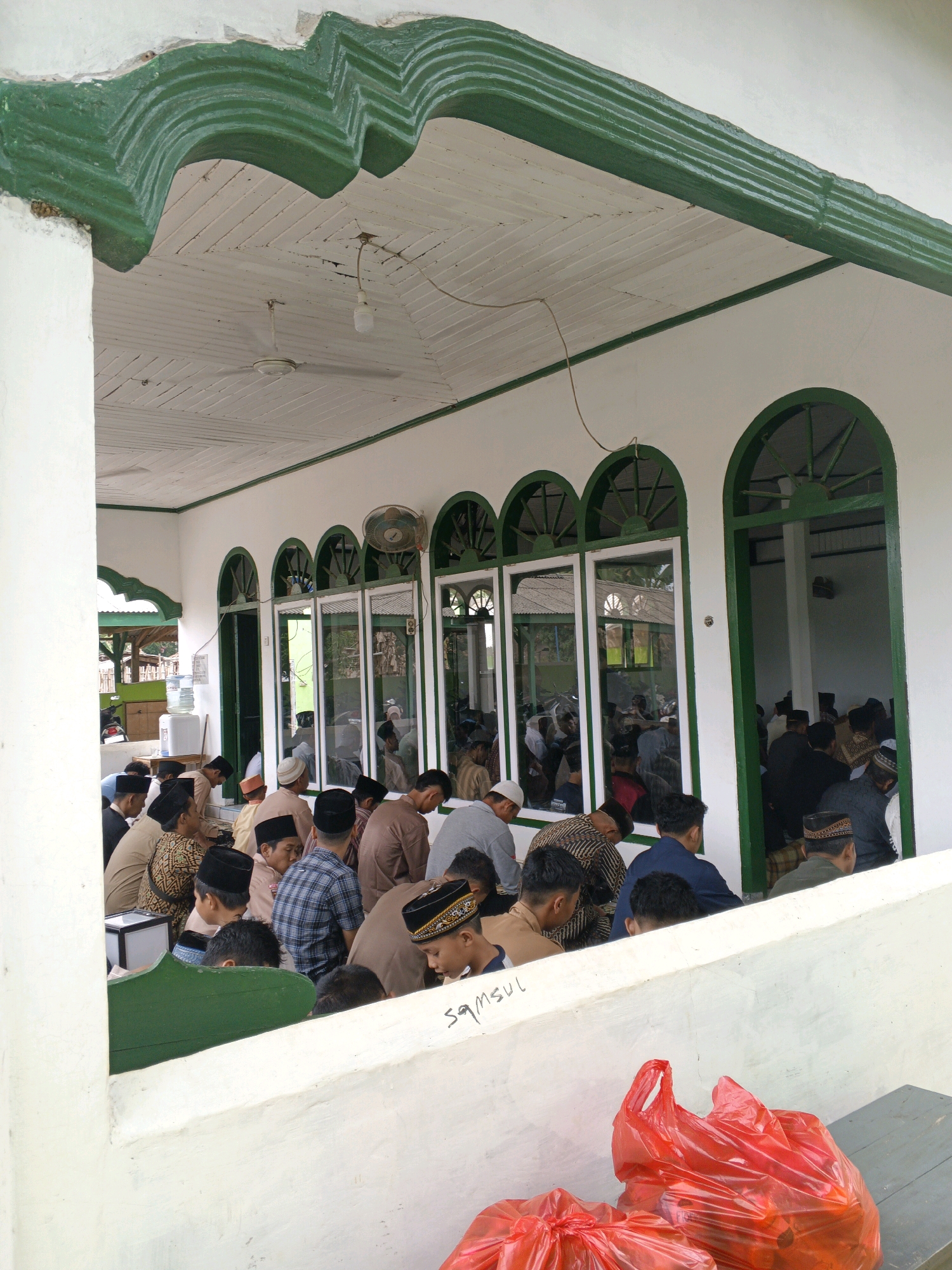 In Gedung Nyapah, Indonesia, a group of Muslim men are gathered for prayer inside a white building with green arched window frames and decorative green trim. Many of the men are wearing peci, a traditional cap, and various forms of Islamic attire. They are seated on the floor, facing a direction that suggests they are praying towards the Qibla. Some men are leaning forward, others are in a sitting position, and some appear to be standing in the background. Reflections in the windows show more people and the interior of the building, indicating a communal prayer service. The environment appears to be a mosque or a similar religious gathering place. Outside the immediate prayer area, a water cooler and a motorcycle are visible through an open doorway, suggesting the location is somewhat rural or semi-urban. In the foreground, two bright red plastic bags are placed on a surface, adding a touch of color to the scene. The overall atmosphere suggests a religious congregation and community gathering.