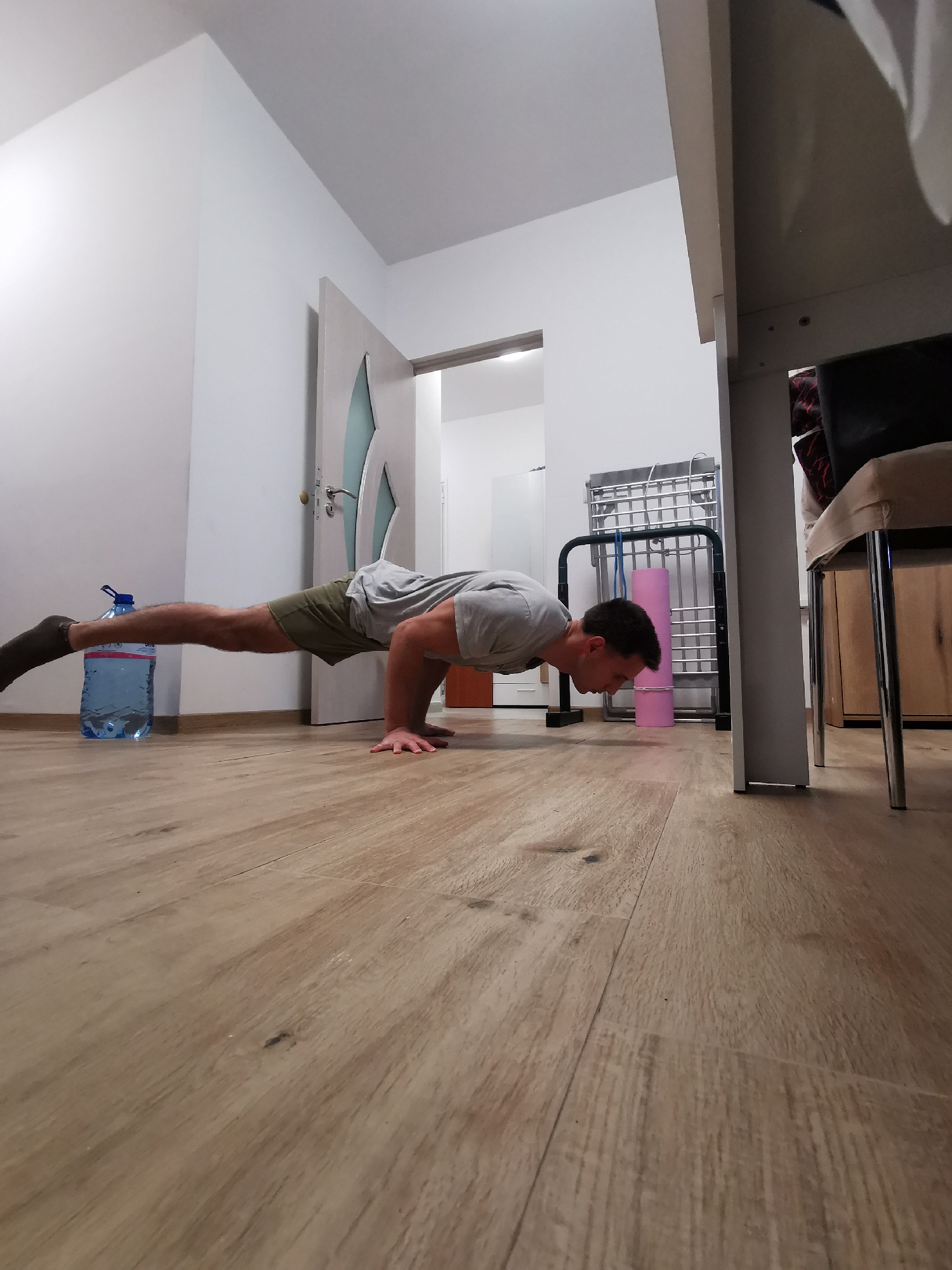 The image displays an indoor scene in Burgas, Bulgaria, featuring a male performing a calisthenics exercise. The male, attired in a grey short-sleeved t-shirt, olive-green shorts, and dark socks, is positioned in a horizontal plank, his torso lowered towards the floor, indicating a push-up or similar variant. His hands are flat on the light-colored laminate wood floor, fingers spread, with his head tilted downwards. His feet are elevated and rest upon a large, clear blue plastic water bottle, which bears a red and white label.
The immediate environment consists of a light-colored laminate wood floor with visible plank seams and a few dark spots, and plain white walls. In the mid-ground, a light-colored interior door with a decorative frosted glass panel and a brass-colored doorknob is partially ajar, revealing a segment of another room or hallway beyond. To the right of the door, a metallic clothing drying rack is visible, alongside a vertically oriented pink yoga mat or foam roller, and a dark piece of exercise equipment on the floor. A portion of a white wardrobe or cabinet is visible on the far left. In the foreground to the right, a section of a furniture item, possibly a table or desk with a light-colored top and dark underside, is present, accompanied by a chair with a light fabric cover and chrome legs. The room is illuminated by ambient light, with a bright light source evident on the upper left wall.