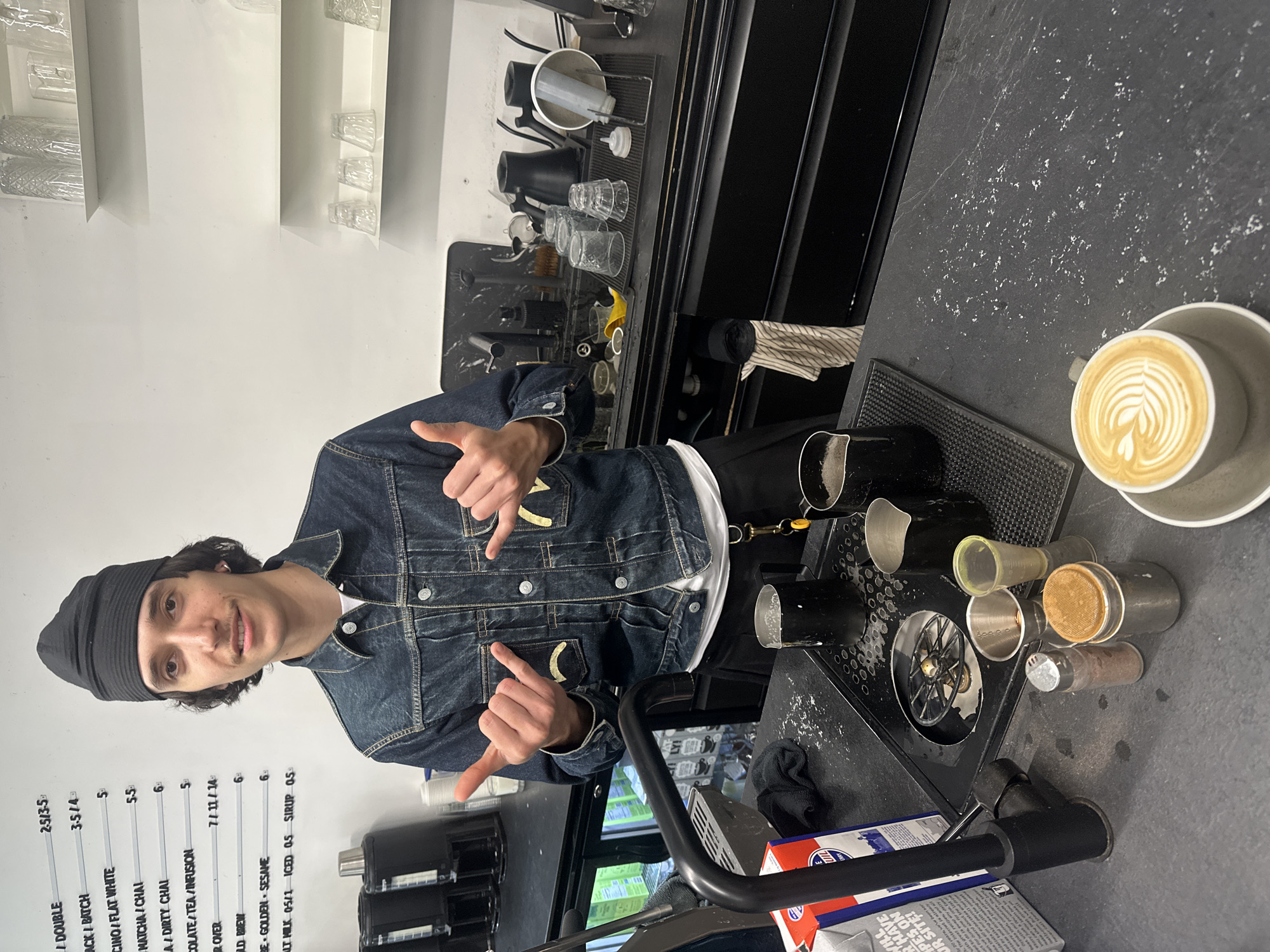 A medium shot depicts a young adult male in a denim jacket and black beanie, positioned in what appears to be a coffee shop. He is making a "hang loose" gesture with both hands, facing the camera. To his right, a countertop is visible with barista tools and a latte cup displaying latte art. Behind him, a menu board listing coffee options with prices is mounted on the wall. The backdrop includes shelves filled with glassware and a professional coffee machine setup. The environment suggests an urban, possibly Parisian, coffee shop setting.