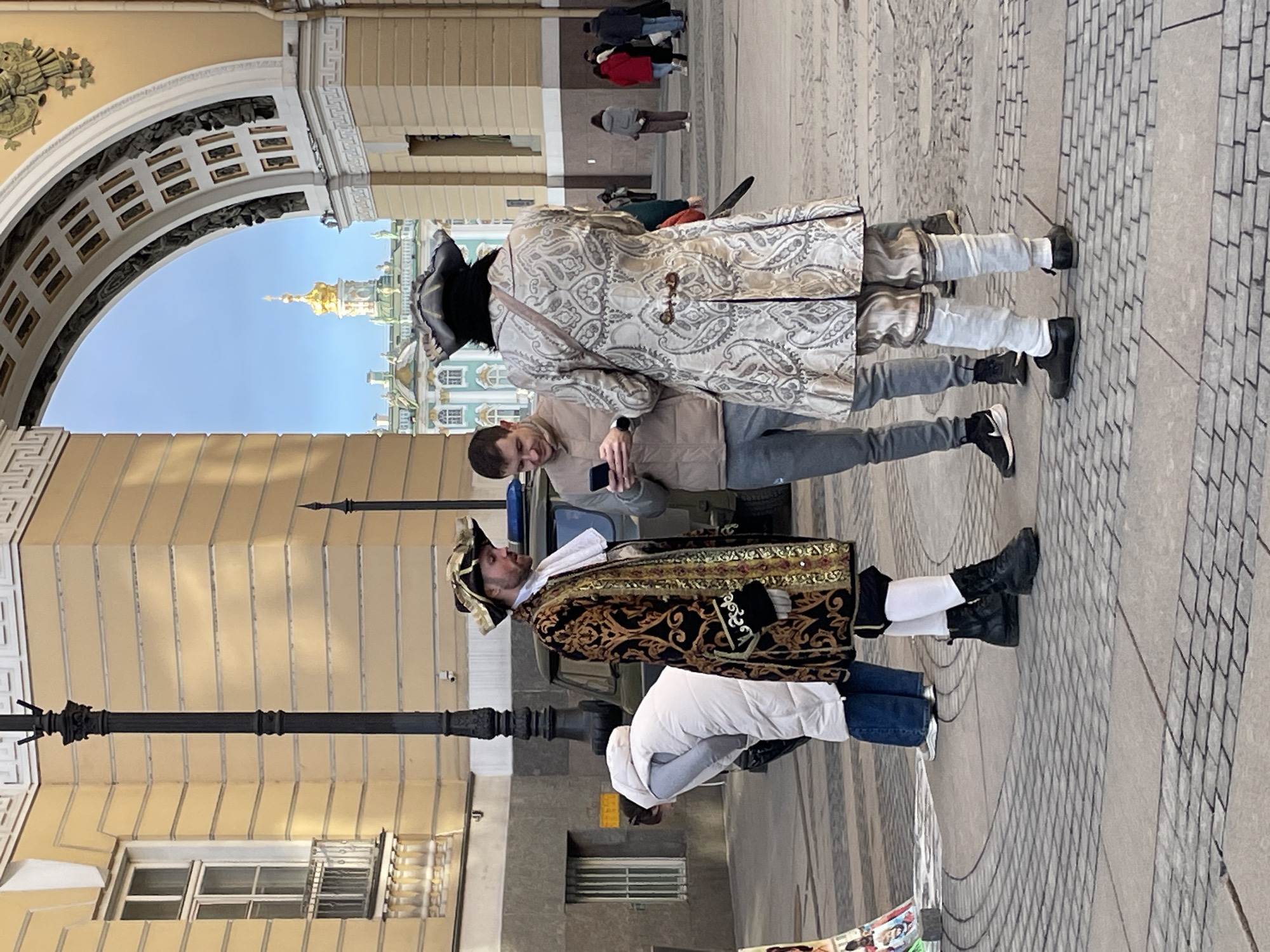 The scene is located in Palace Square, Saint Petersburg, Russia, beneath the Arch of the General Staff Building, with the Winter Palace visible through the archway in the background.
Three adult male figures are positioned in the foreground on a paved surface. To the left, a man with a beard wears a dark tricorne hat, a black and gold brocade long coat with a white cravat, white breeches, and black combat boots. He is looking towards a smartphone. Centrally, another man in modern attire, wearing a light brown puffer vest over a long-sleeved shirt, grey jeans, and black sneakers, holds a black smartphone and appears to be actively viewing its screen. To the right, a third man, observed from the back, wears a dark tricorne hat, a light-colored, patterned long coat with pleats, light breeches, and dark flat shoes. A dark strap crosses his back, and a dark handle, possibly of a prop sword, protrudes from his left side.
In the mid-ground, behind the central figures, a dark green utility vehicle, possibly a UAZ model, is parked. A tall, black ornamental lamppost stands between the left-most costumed man and the vehicle. To the left of the lamppost, a woman in a white puffer jacket and blue jeans is bent over, facing away from the camera. A colorful vertical display stand is partially visible near her. In the background, beyond the immediate square, multiple indistinct figures are walking. The surrounding architecture consists of large, light yellow buildings with white trim and window frames. The blue sky is visible through the archway, framing the distinct golden dome of the Winter Palace. The ground is paved with grey interlocking pavers.